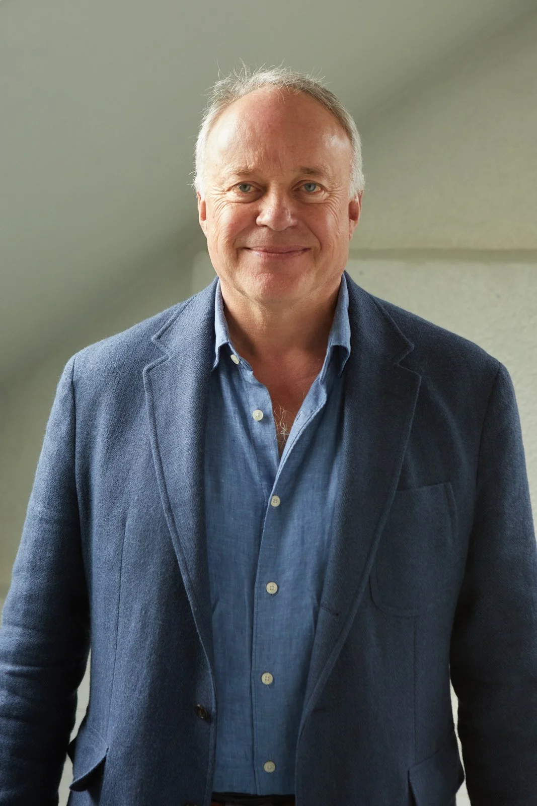 A smiling middle-aged man with light-colored hair, wearing a blue shirt and a blue blazer, standing indoors against a plain wall.