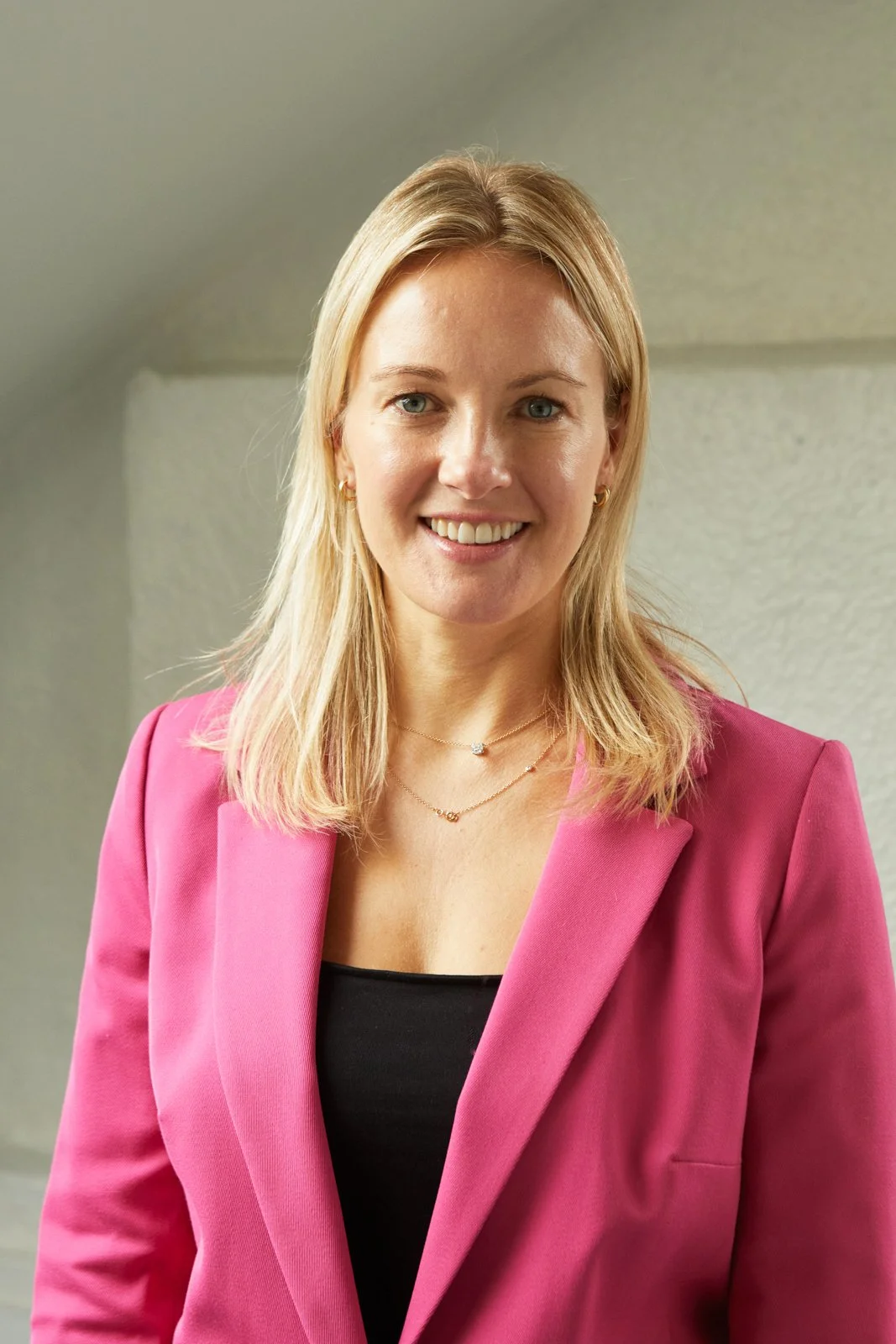 A smiling woman with blonde hair wearing a pink blazer and a black top, standing indoors against a neutral background.