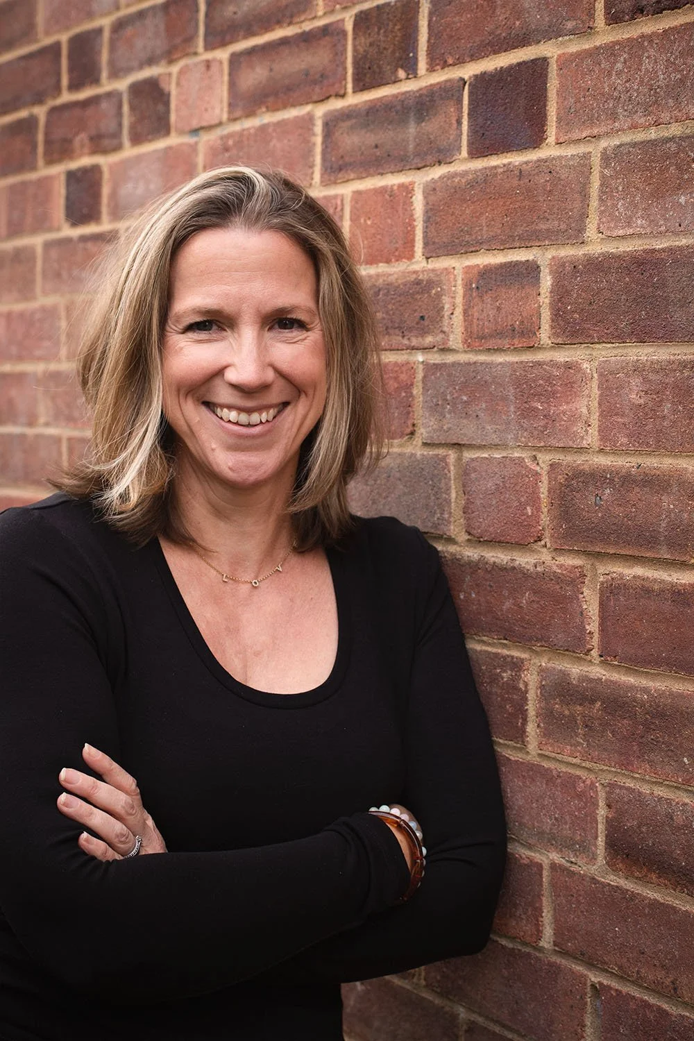A woman with shoulder-length blonde hair smiling, standing with arms crossed, wearing a black top, against a red brick wall.