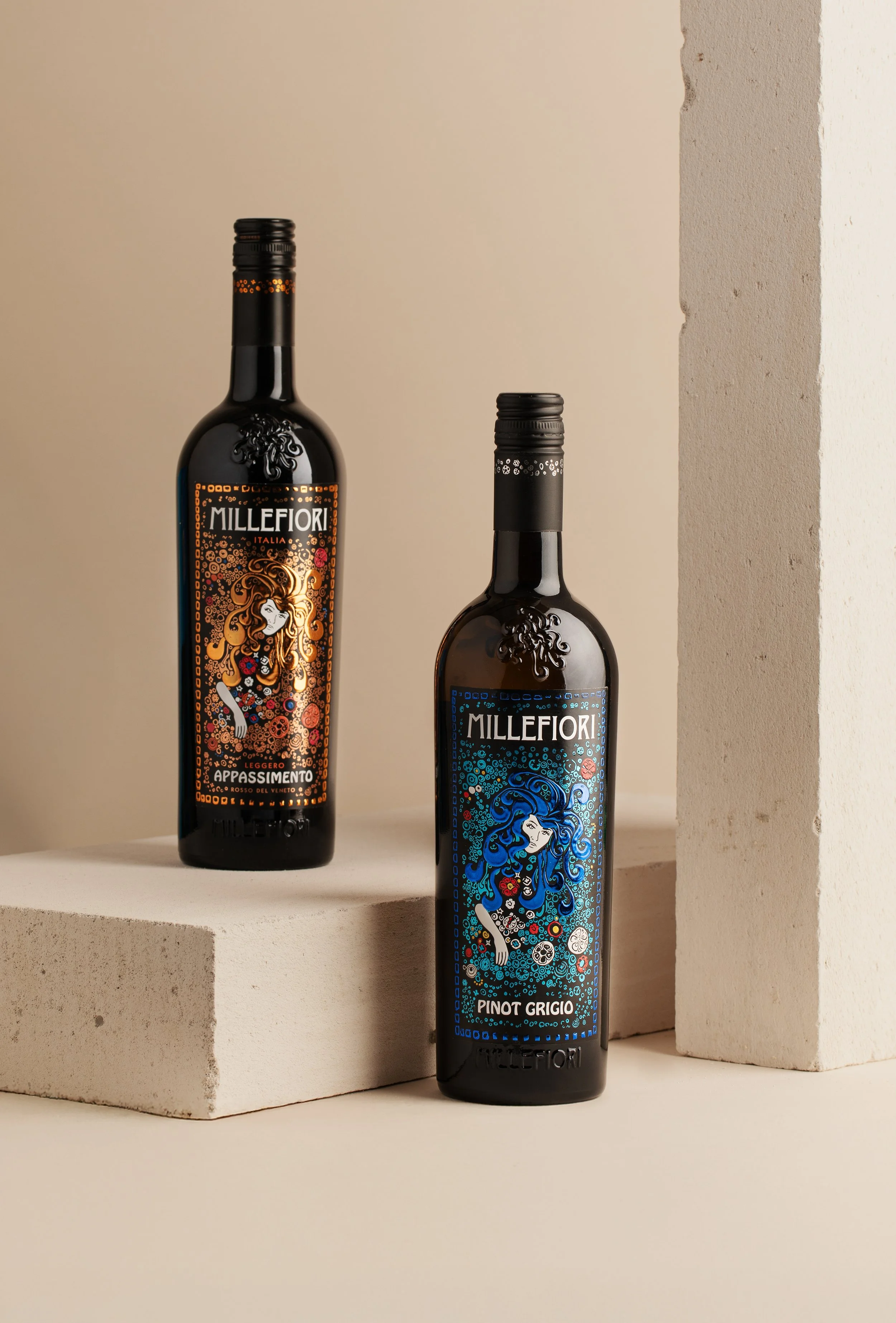 Two bottles of Millefiori wine, one with orange label and the other with blue label, featuring artistic illustrations of women with flowing hair surrounded by patterns, set against minimalist background with neutral-colored blocks.