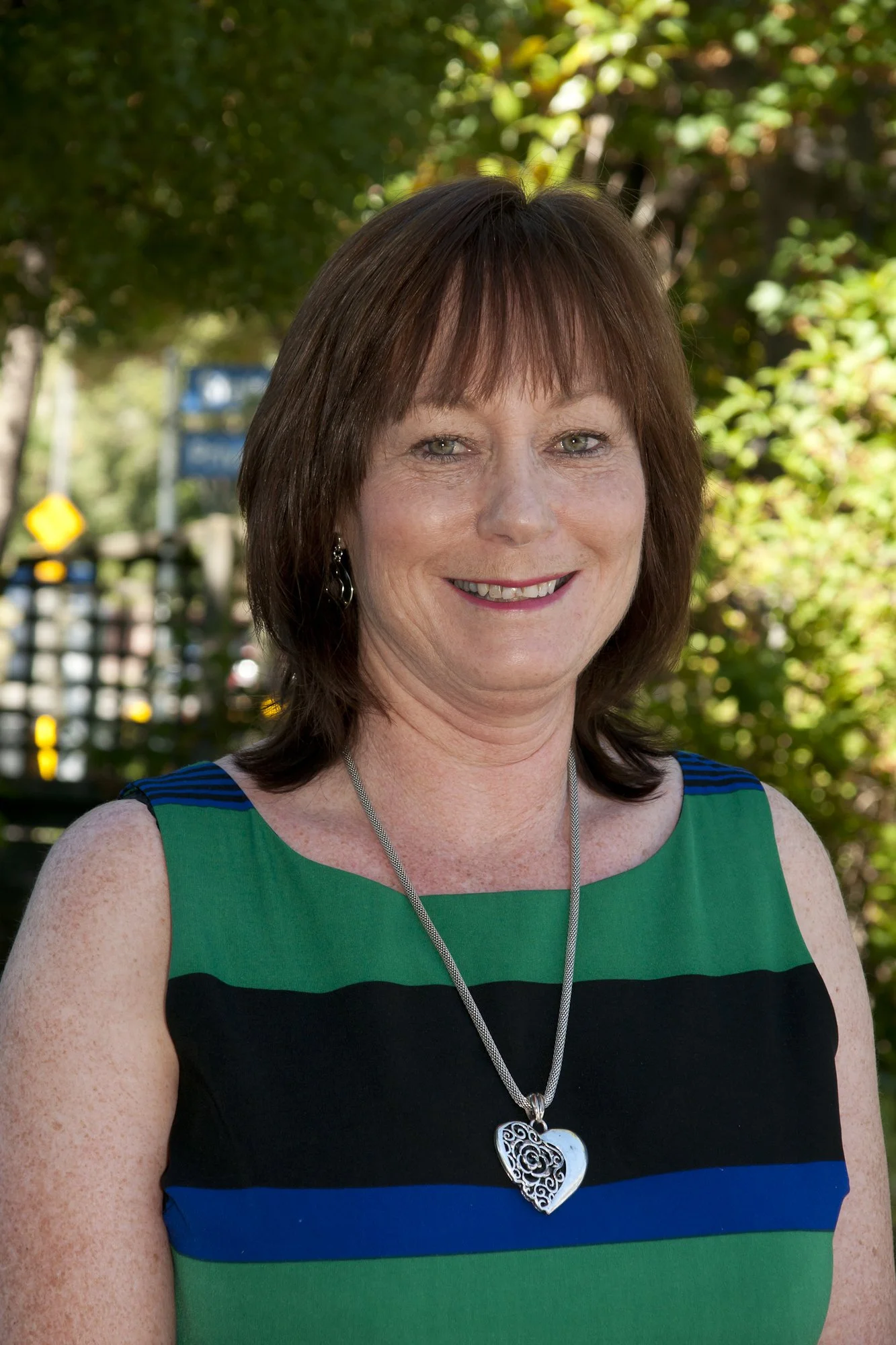 A woman with shoulder-length brown hair, wearing a sleeveless dress with green, black, blue, and teal horizontal stripes, standing outdoors with green trees and a partly sunny sky in the background.