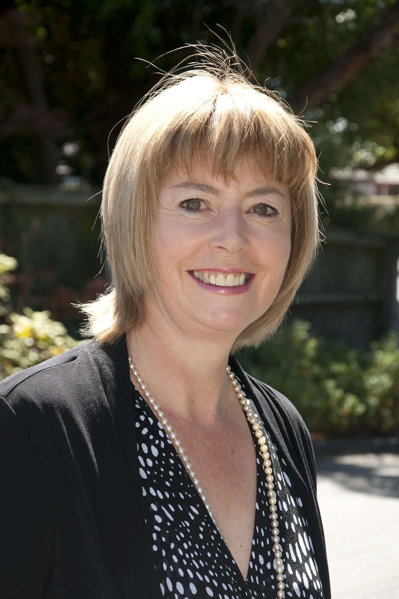 A woman with short blonde hair smiling outdoors on a sunny day, wearing a black jacket, a black and white patterned top, and a pearl necklace.