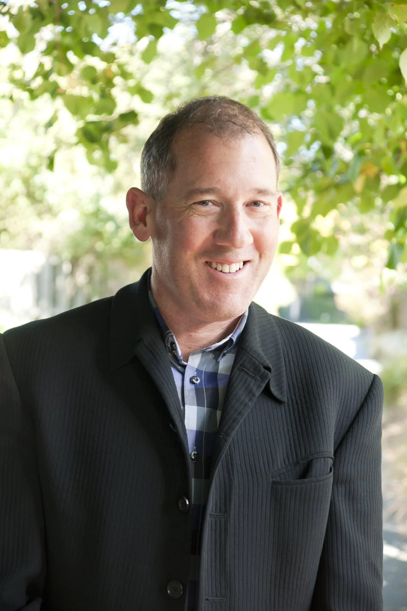A smiling man in a black suit jacket and checkered shirt standing outdoors with green foliage in the background.