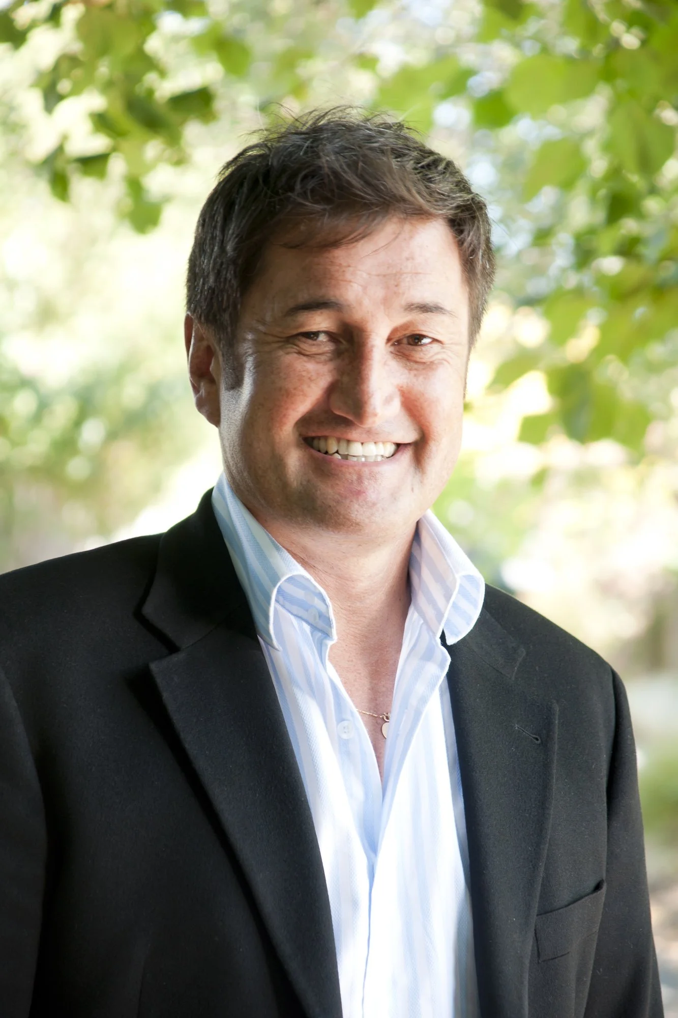 A smiling man in a suit with a light blue striped shirt, standing outdoors with green foliage in the background.