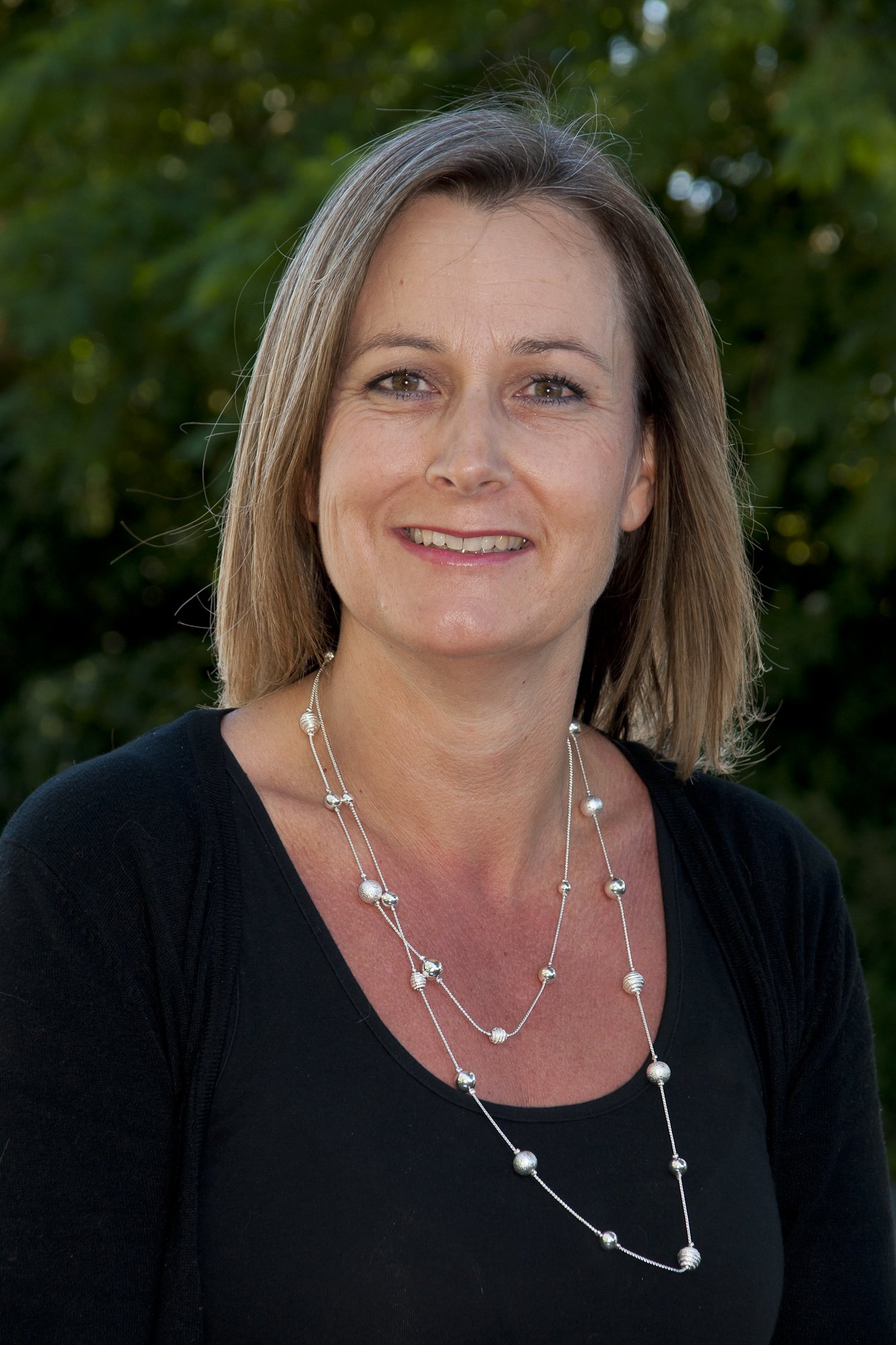 A woman with shoulder-length blonde hair, smiling, wearing a black top and silver beaded necklaces, standing outdoors with a background of green trees.