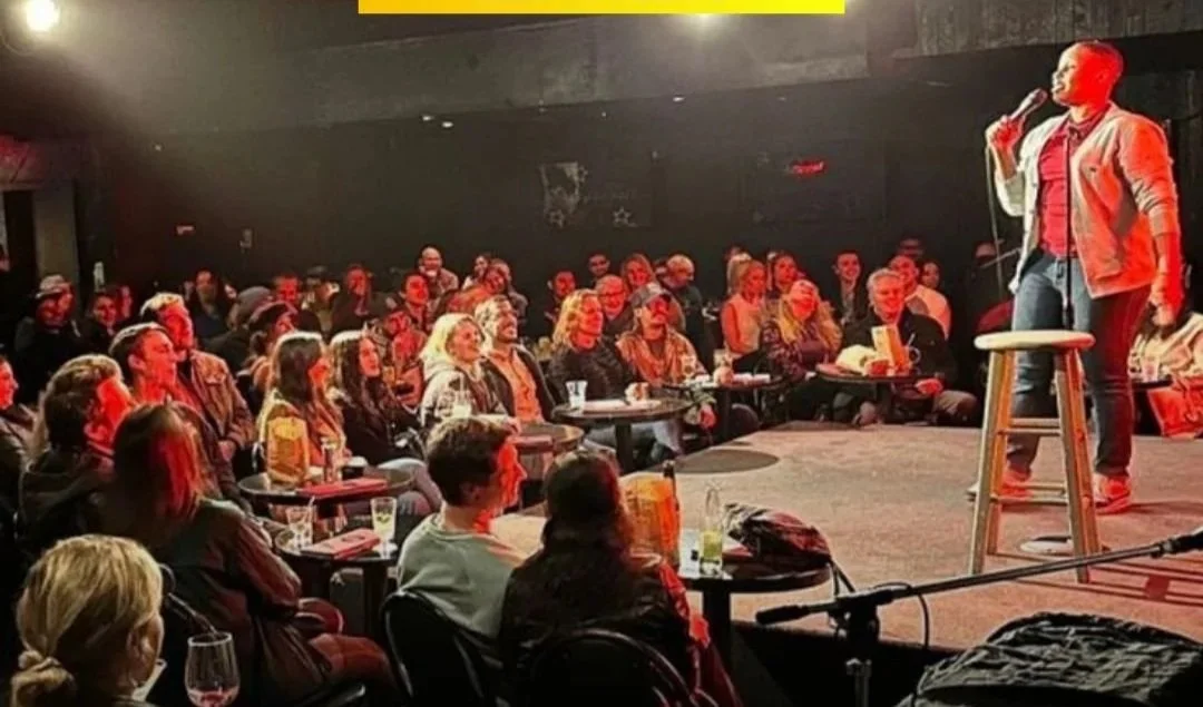 A stand-up comedy show with a female comedian performing on stage, holding a microphone and a stool, in front of an audience seated at tables.