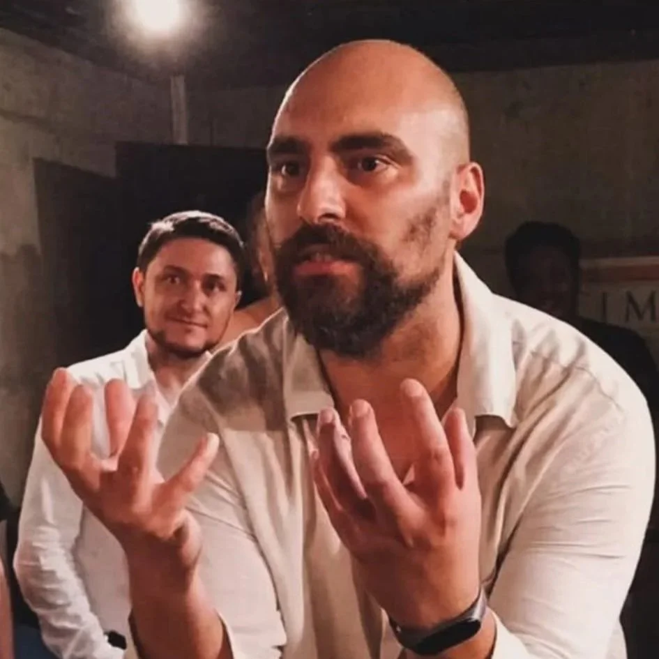 Two men, one with a beard and bald head, gesturing with crossed hands in a dimly lit indoor setting.