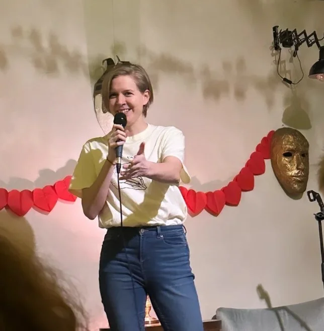 A woman smiling and holding a microphone on a stage decorated with a red paper heart chain and a gold mask on the wall behind her.