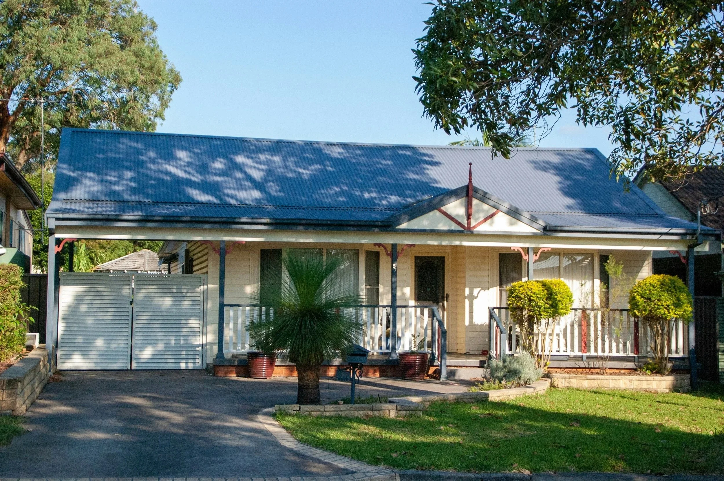 Front view of a house with a blue corrugated metal roof, white exterior walls, and a porch. There are plants and trees in the yard, including a palm in a pot and shaded bushes. The driveway and sidewalk are visible in the foreground, with sunlight filtering through the trees.