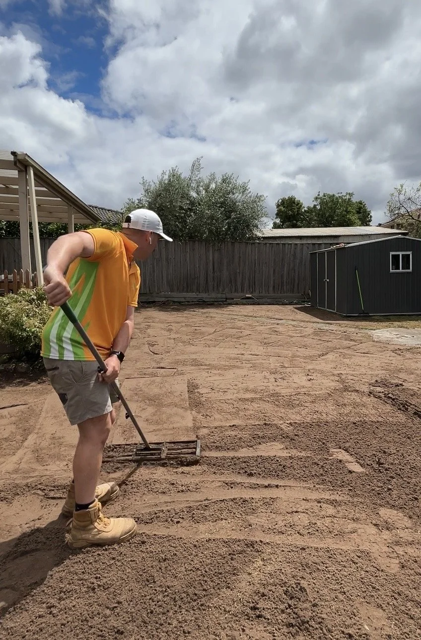A man in a yellow shirt, beige shorts, and tan work boots is raking a large area of dirt in a backyard. He is wearing a white cap and black watch. The backyard has a wooden fence, some trees, a small shed, and a partly cloudy sky.