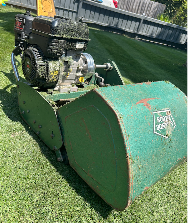 A green Scott Bonar lawn roller with a black gas-powered engine on a neatly cut grass lawn.