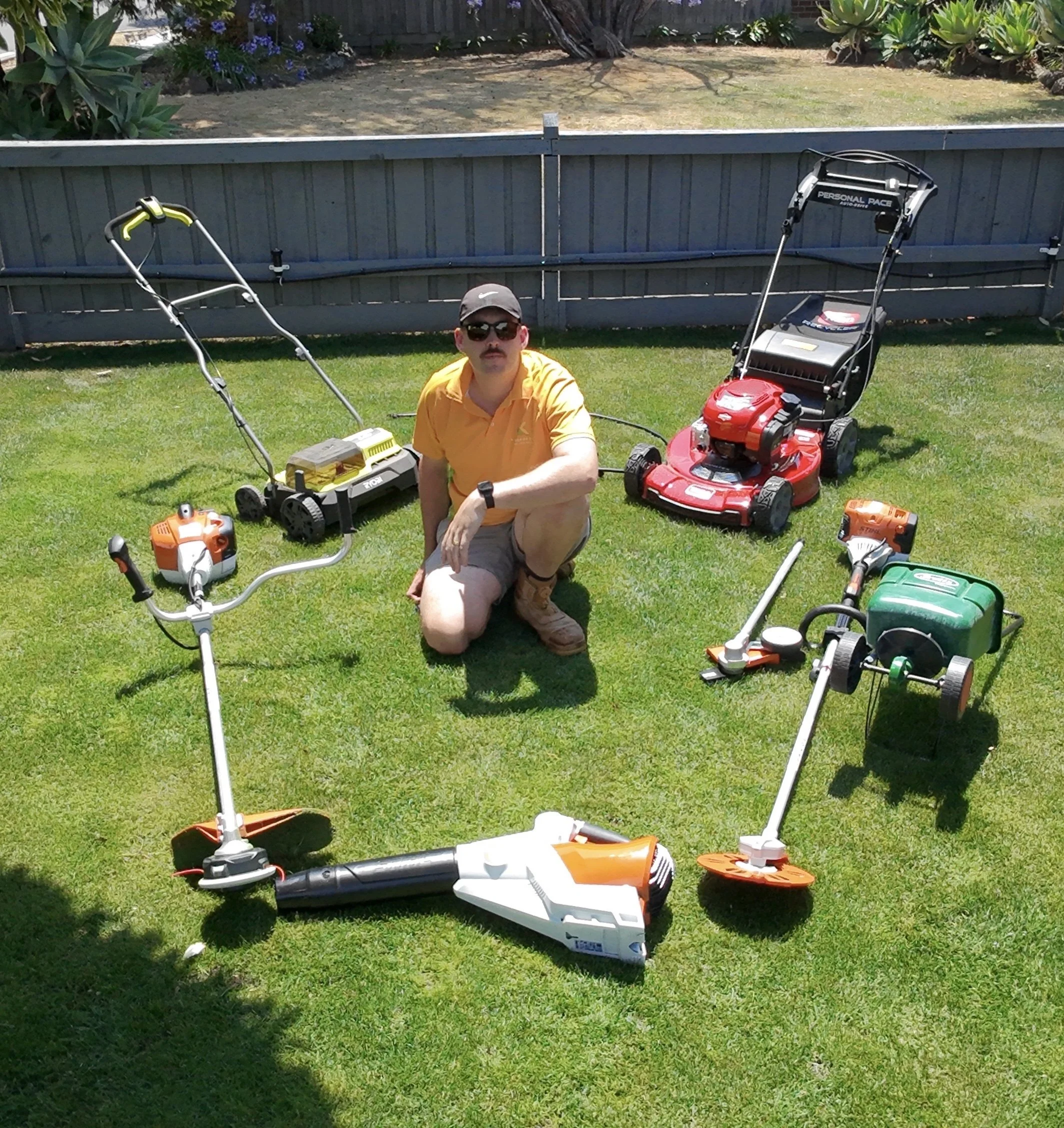 Man kneeling on a green lawn surrounded by various lawn care equipment including a push mower, string trimmer, leaf blower, and other yard tools. There is a fence and some plants and trees in the background.