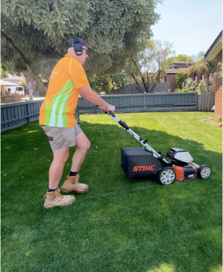 Person wearing an orange and green shirt, shorts, and boots mowing the lawn with a gas-powered lawn mower in a backyard with trees and a fence.