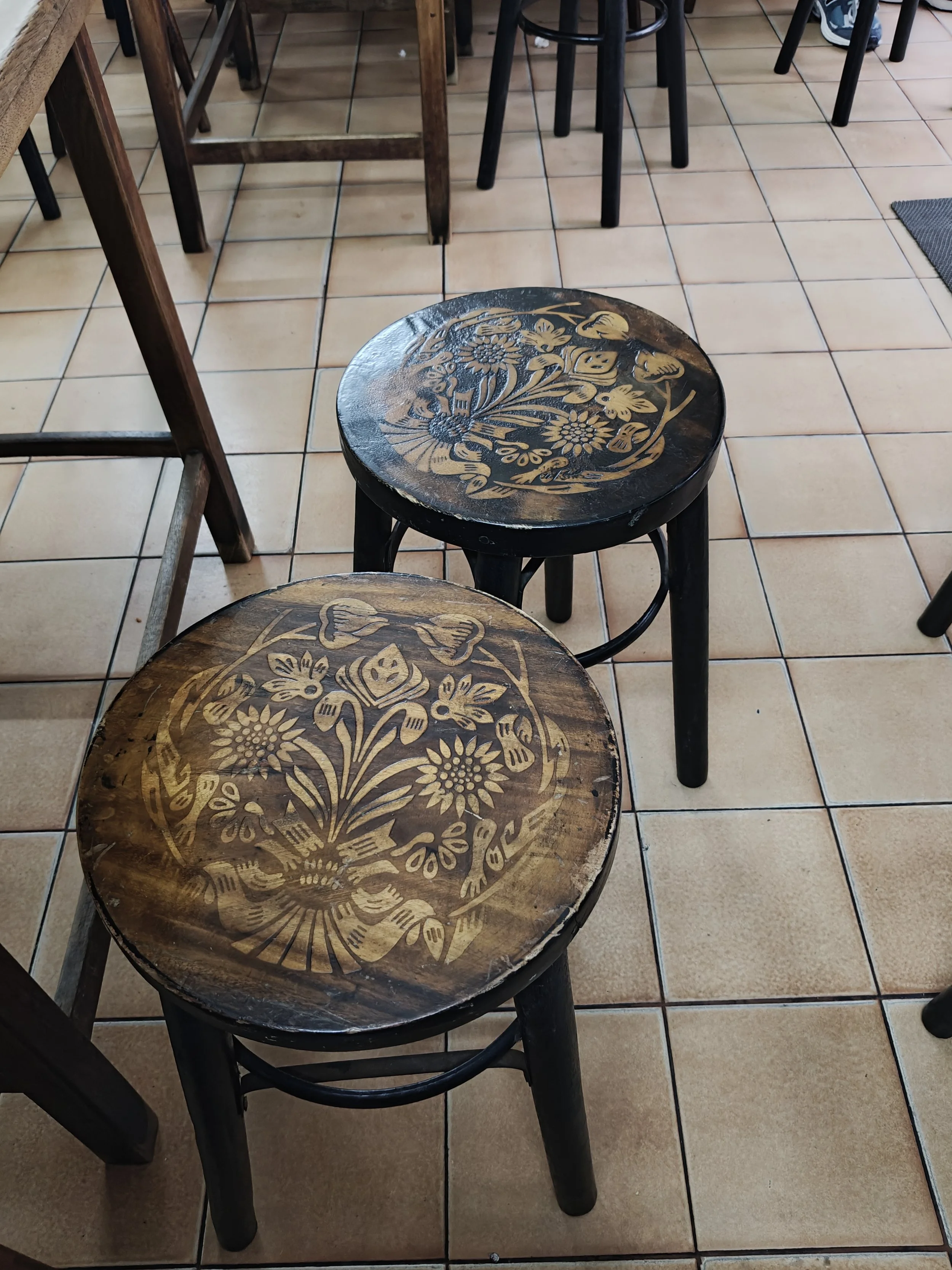 Two wooden stools with floral designs on the tops, placed on a tiled floor in a restaurant or cafe setting.