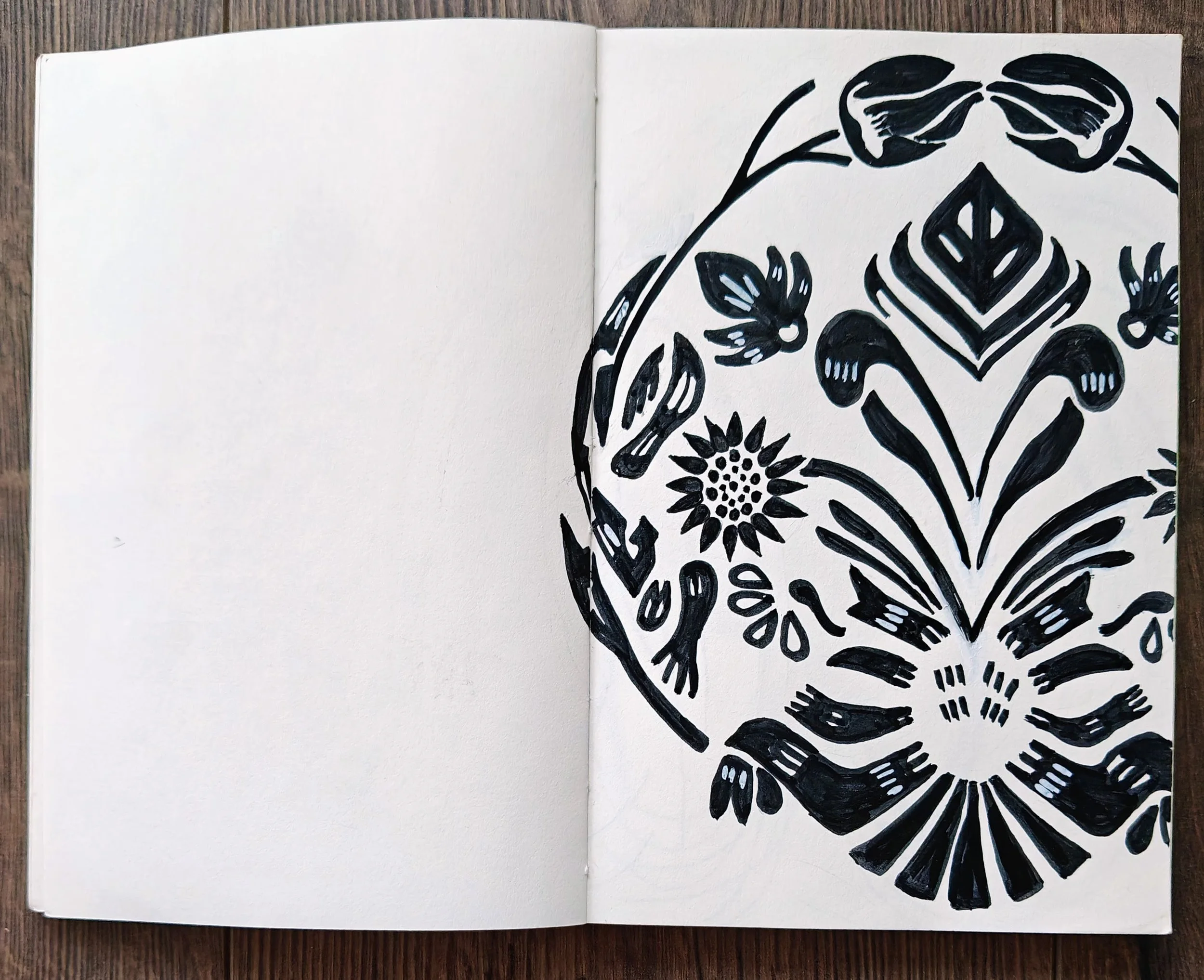 Open sketchbook with black abstract floral and geometric designs on the right page, left page blank, placed on a wooden surface.