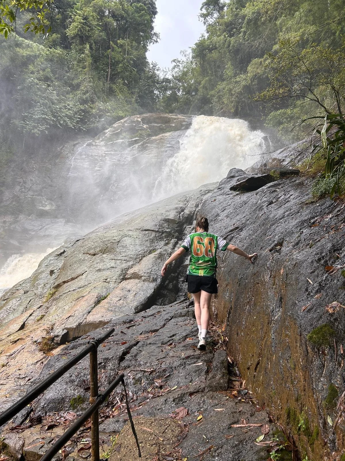 Person wearing a green sports jersey and black shorts, walking on a wet rocky trail beside a waterfall in a lush, forested area.
