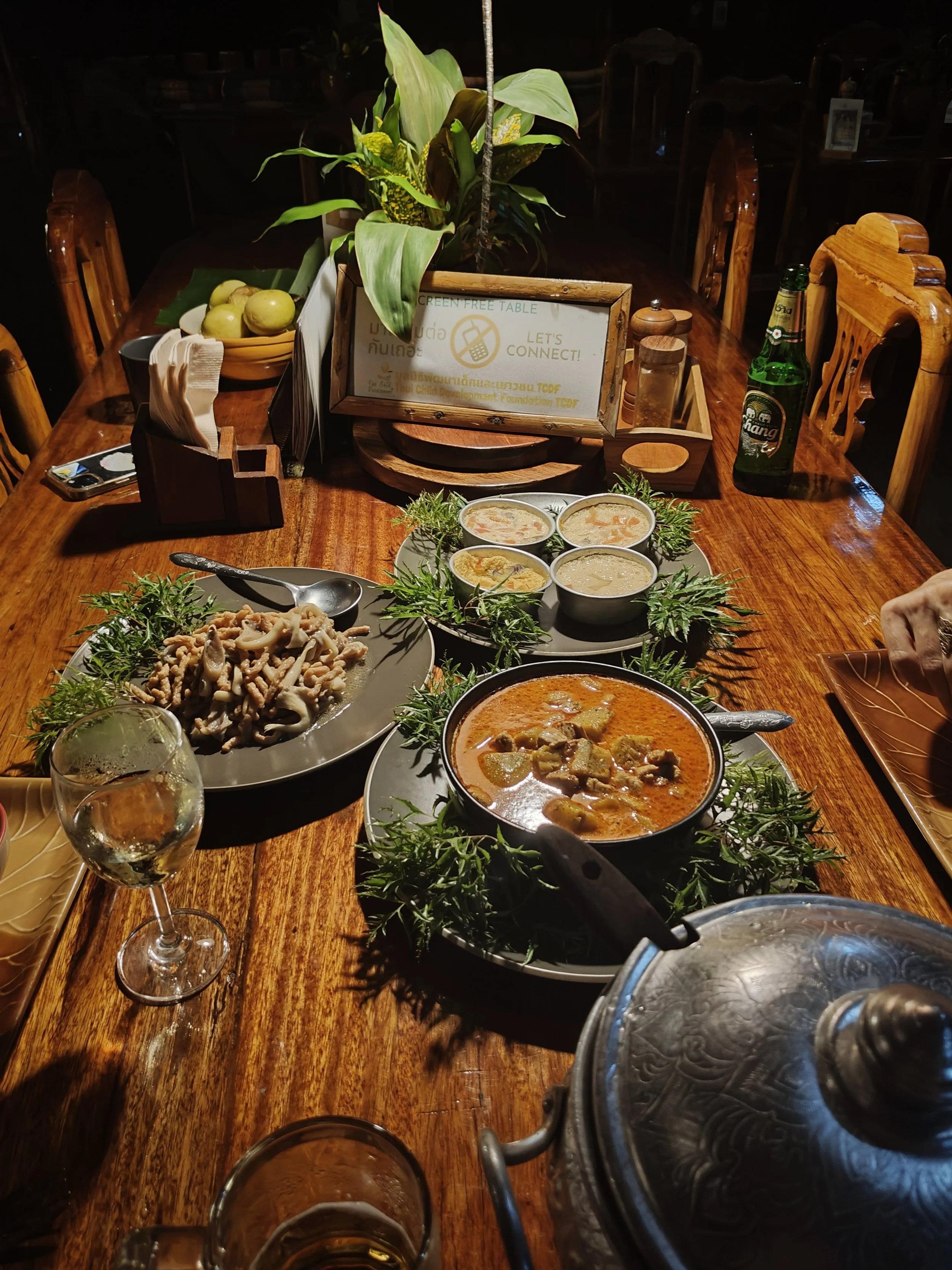 A wooden dining table set with various Thai dishes, including a curry, a plate of stir-fried mushrooms, and four small bowls of soup, surrounded by chairs and condiments. A sign on the table indicates a contactless or green table initiative, and ther