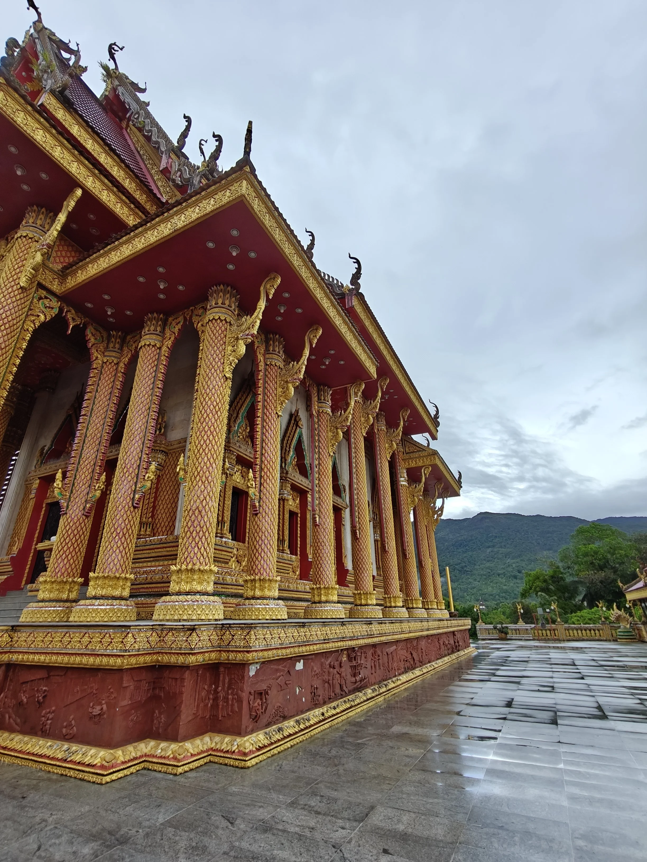 A detailed view of a Thai temple with ornate gold and red decorations, tall columns with intricate designs, and a cloudy sky in the background.