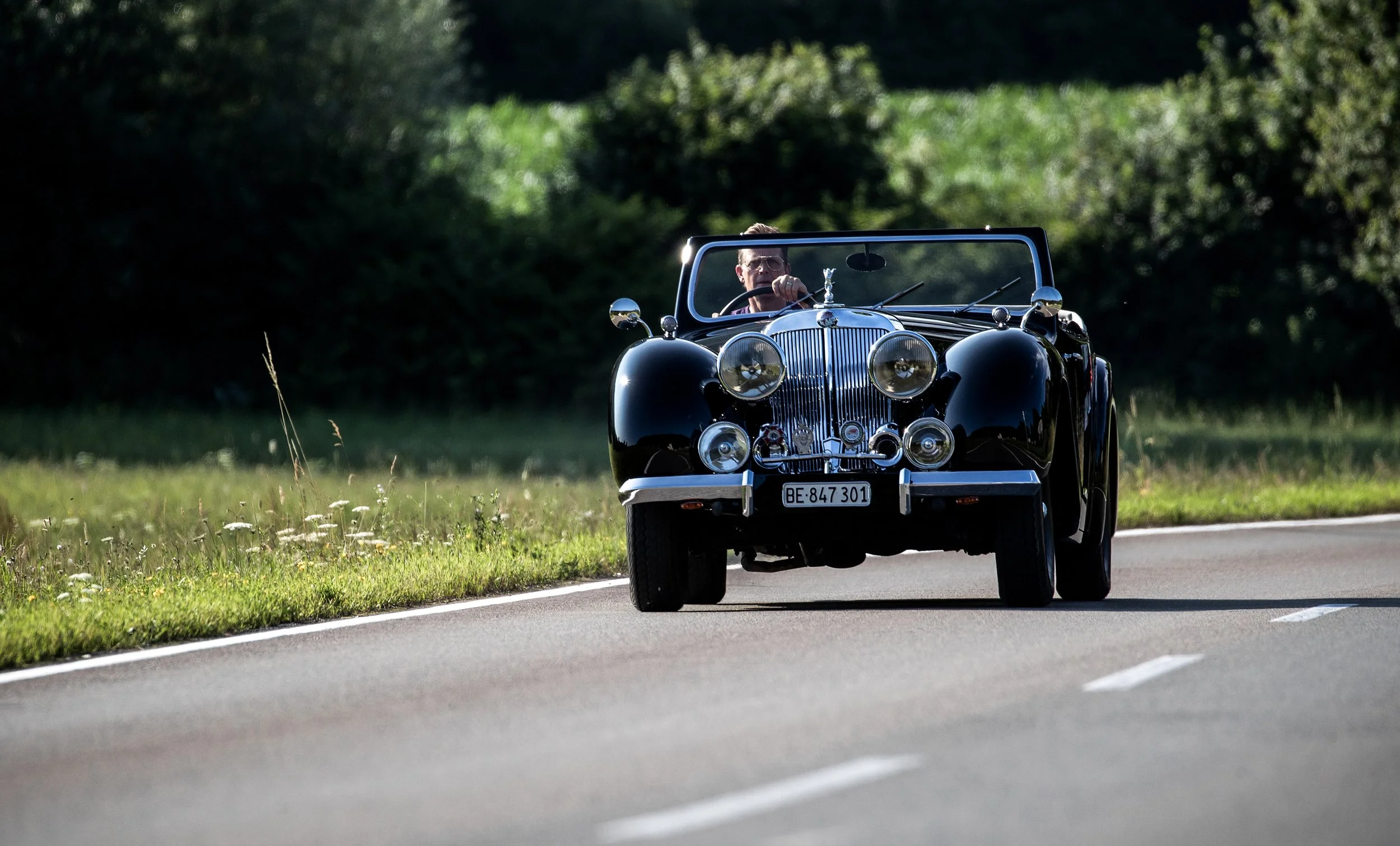 Triumph Roadster 2000 Oldtimer Cabriolet auf Landstraße. Schwarze Lackierung mit klassischer Frontpartie und verchromten Scheinwerfern. Restauriertes britisches Fahrzeug der späten 1940er Jahre im Sonnenlicht.