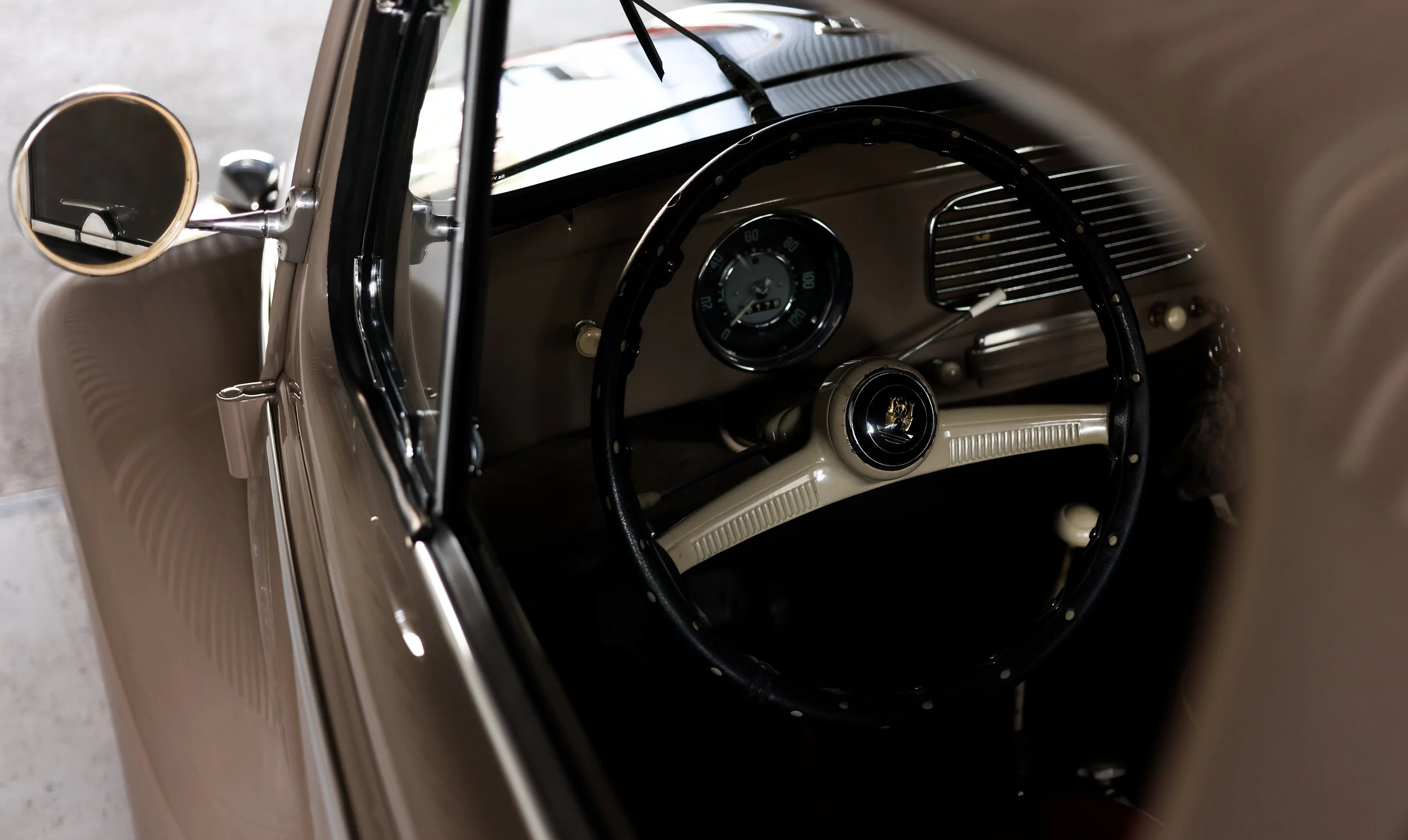 VW Käfer Oldtimer Detailaufnahme des Innenraums. Blick durch das geöffnete Seitenfenster auf das originale Lenkrad und Armaturenbrett. Braun-graue Karosserie mit originalgetreu restauriertem Interieur der 1950er Jahre.