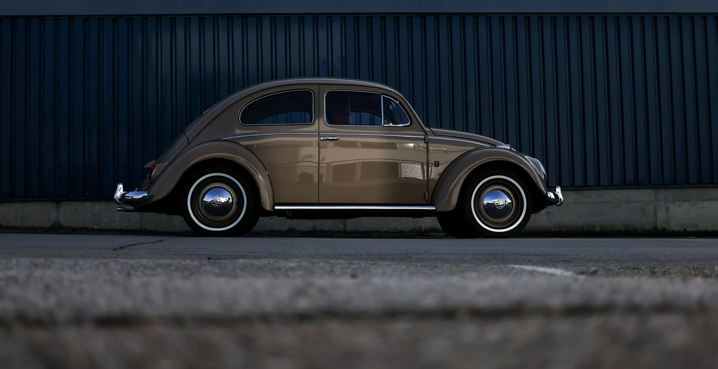 VW Käfer Oldtimer in Seitenansicht vor einer blauen Industriehalle. Braun-graue Lackierung mit originalgetreuer Restaurierung. 