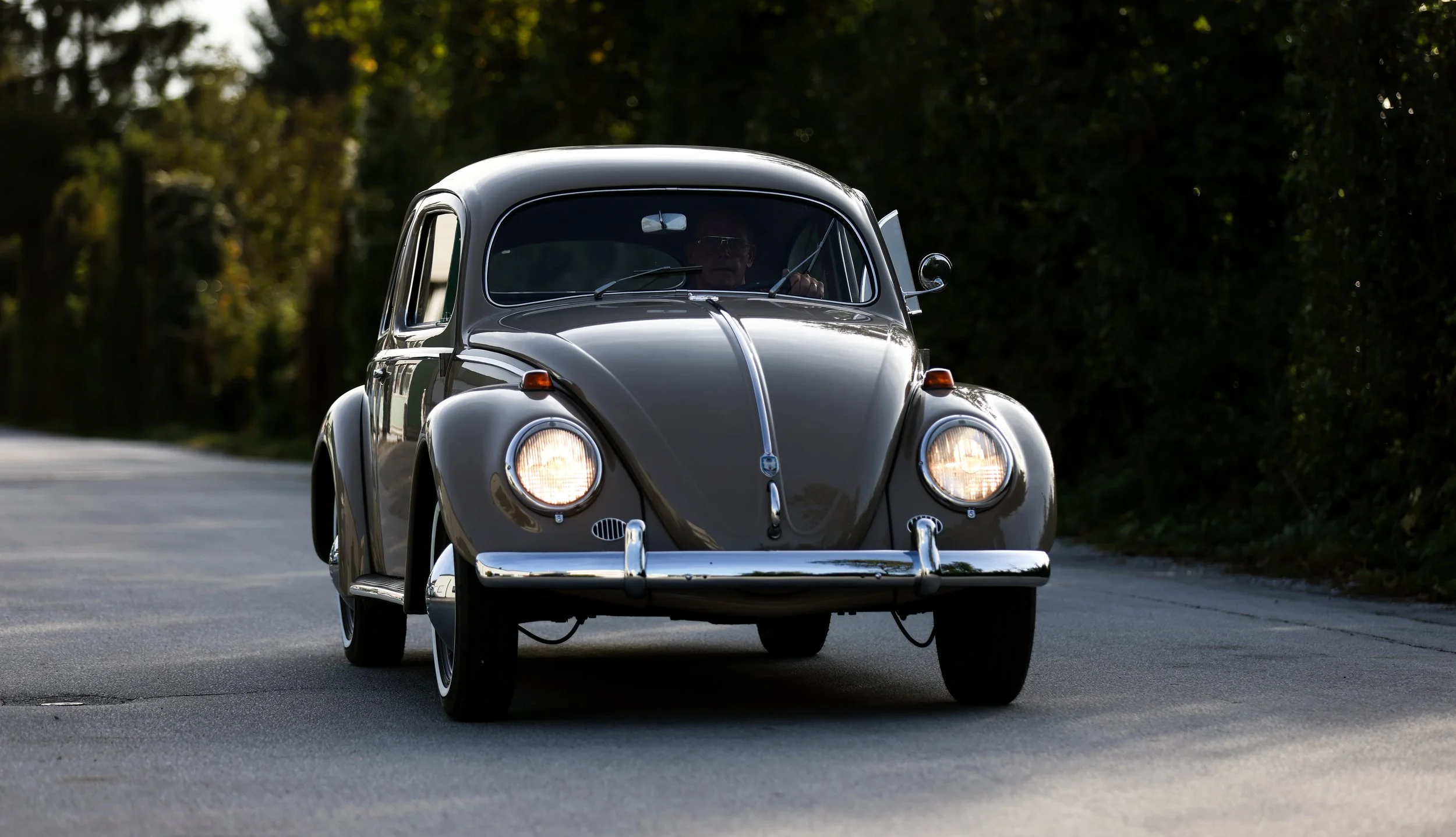 VW Käfer Oldtimer in Fahrt auf einer Landstraße. Originalgetreue Restaurierung mit braun-grauer Lackierung und klassischem Design der 1950er Jahre. Restauriertes Fahrzeug als Beispiel hochwertiger Lackierarbeit.