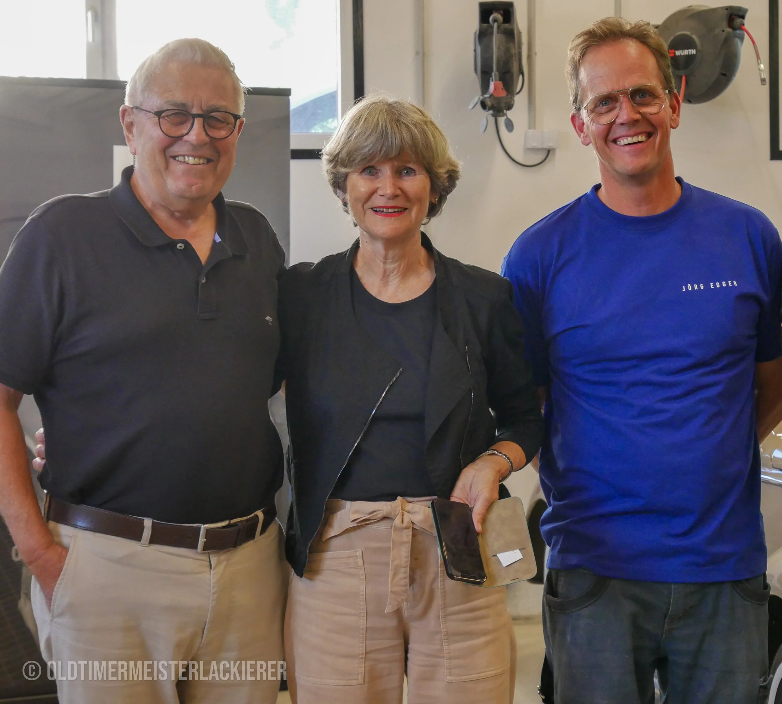 Zwei Kunden eines Triumph Roadster 2000 stehen gemeinsam mit dem Oldtimermeisterlackierer in der Werkstatt in Hallein, Salzburg.