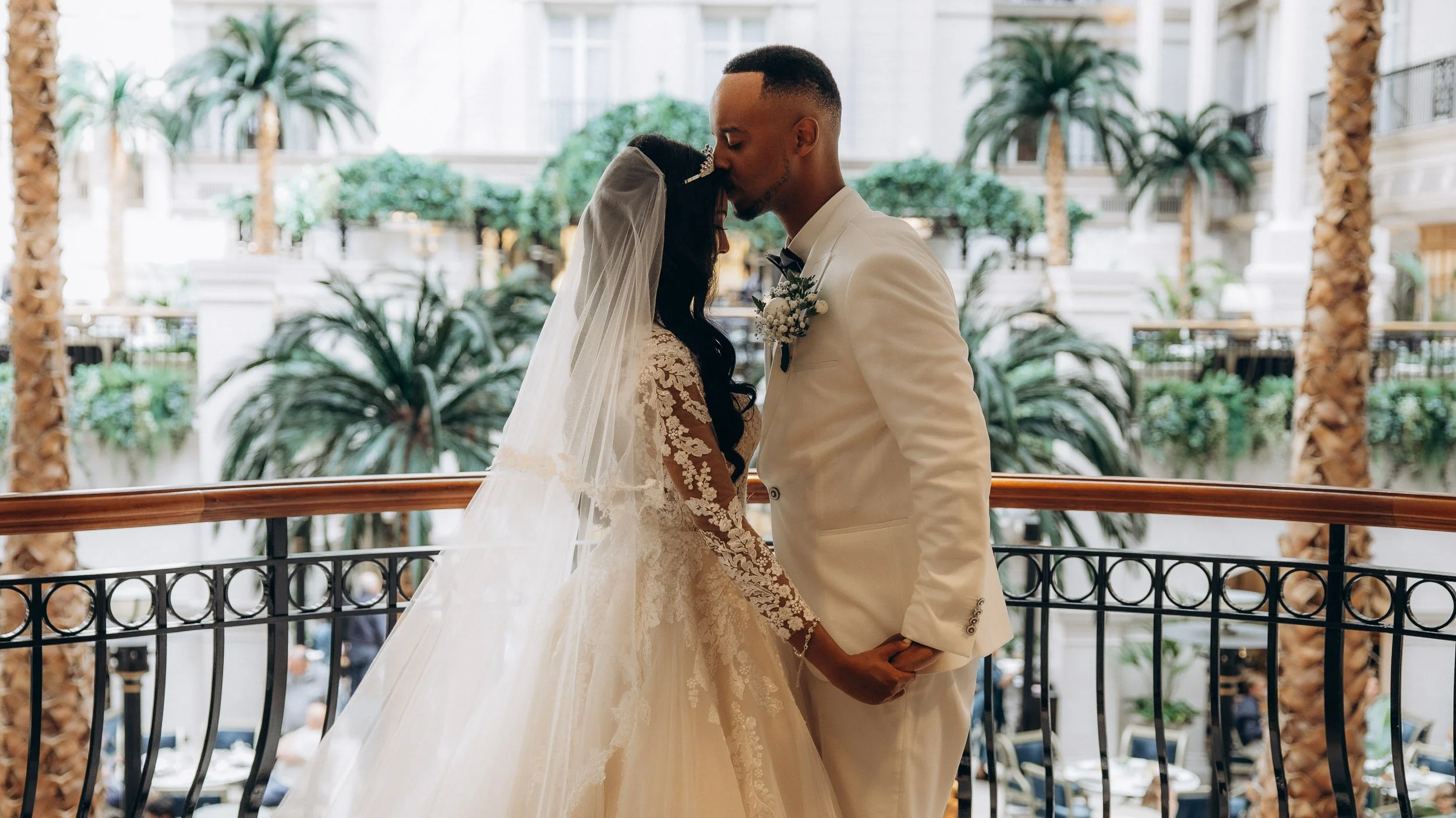 An emotional moment as the newly married couple, bride and groom in white, share a kiss on an ornate indoor balcony overlooking the [Luxury Hotel] atrium; capturing romance and upscale elegance in London wedding photography.
