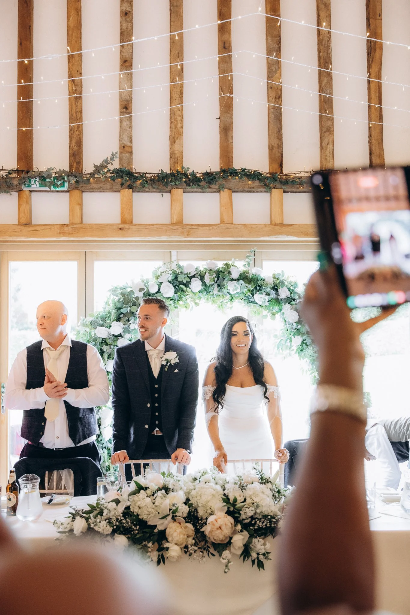 Guest filming the ceremony on a phone as the couple stands beneath a floral arch