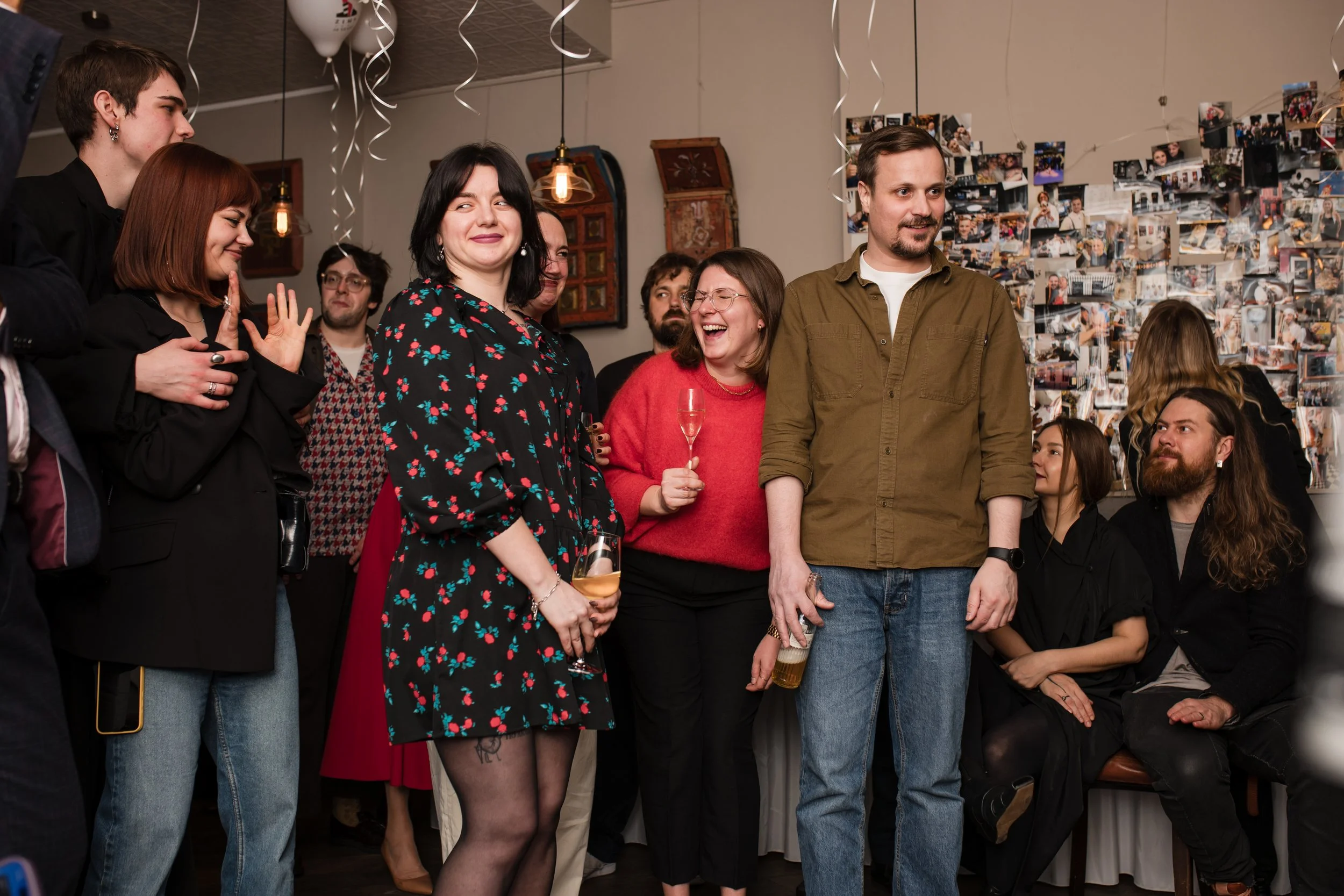 Group of people at a celebration or party, smiling and enjoying themselves. Some are holding drinks, and the background features a collage of photos on the wall.