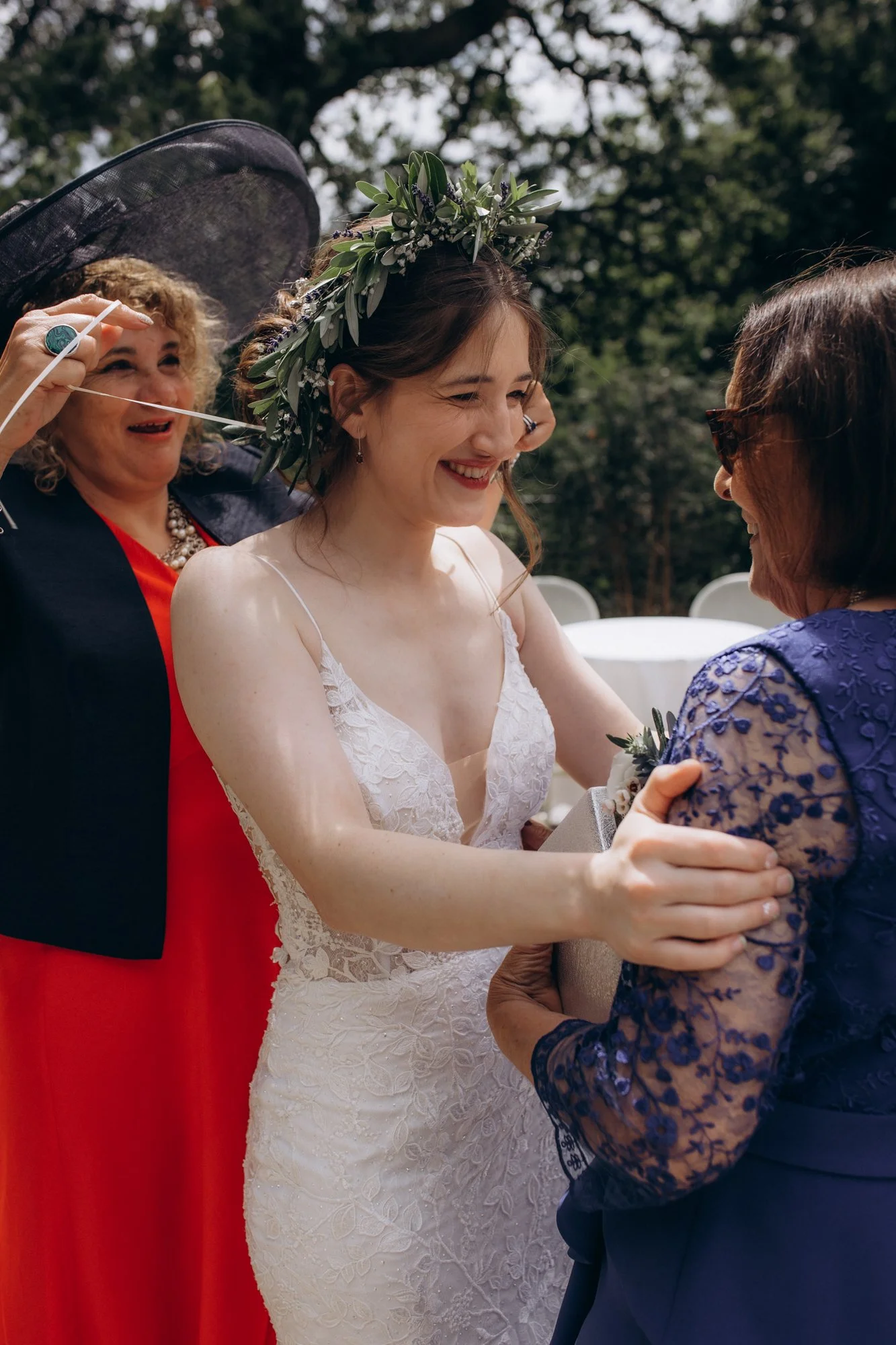A bride in a white lace wedding dress with a floral crown is smiling and holding hands with an older woman in a blue lace dress. An older woman wearing a black hat with a wide brim and a red dress is in the background, smiling.