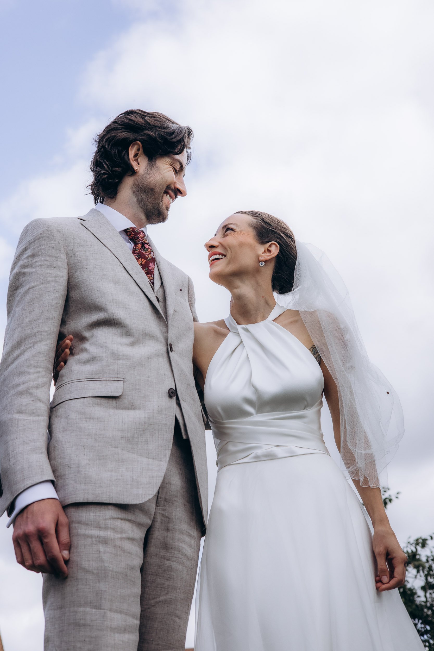  Low-angle portrait of bride and groom gazing at each other and smiling against a bright cloudy sky, bride in a white satin halterneck gown with veil, groom in a light grey suit with a burgundy floral tie 