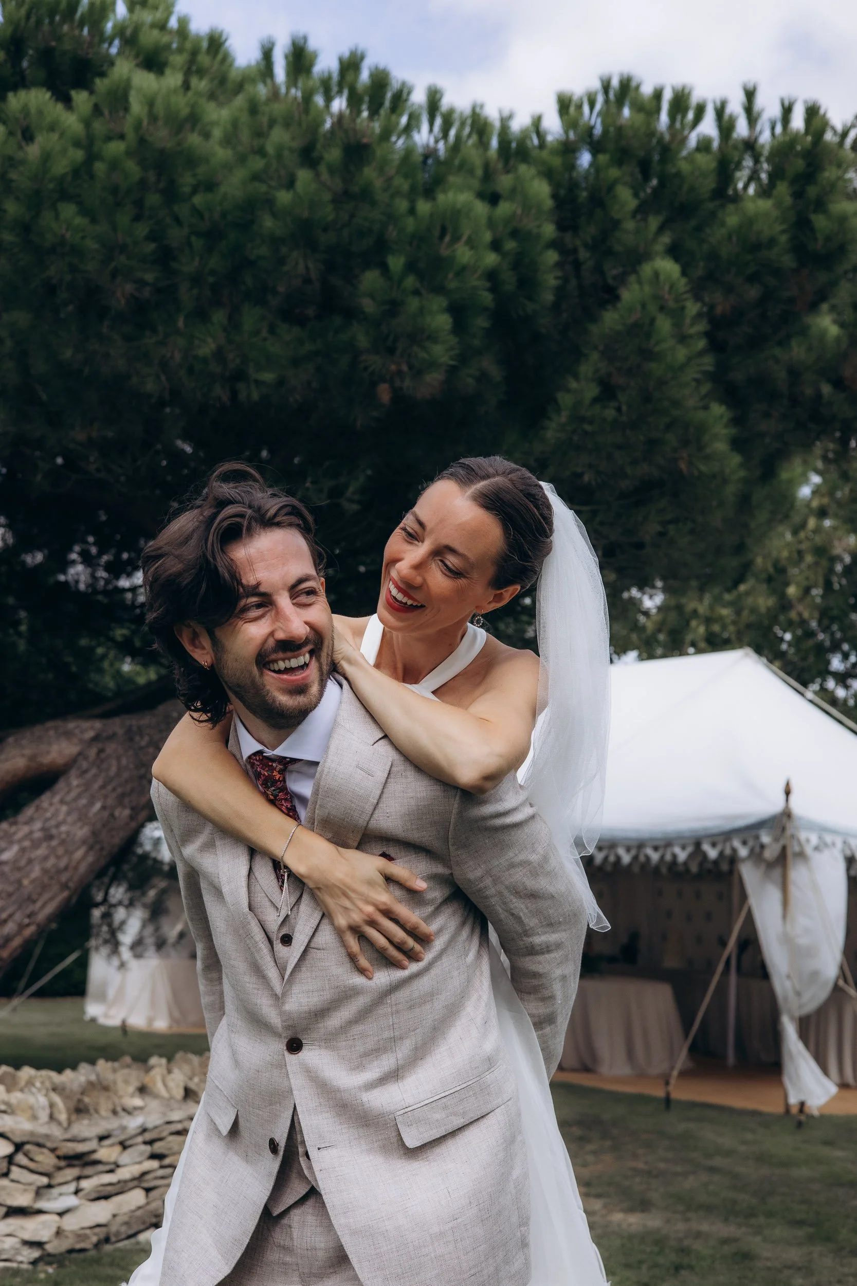  Bride laughing and wrapping her arms around the groom's neck from behind in a playful piggyback pose in a garden, with a white marquee tent visible in the background 