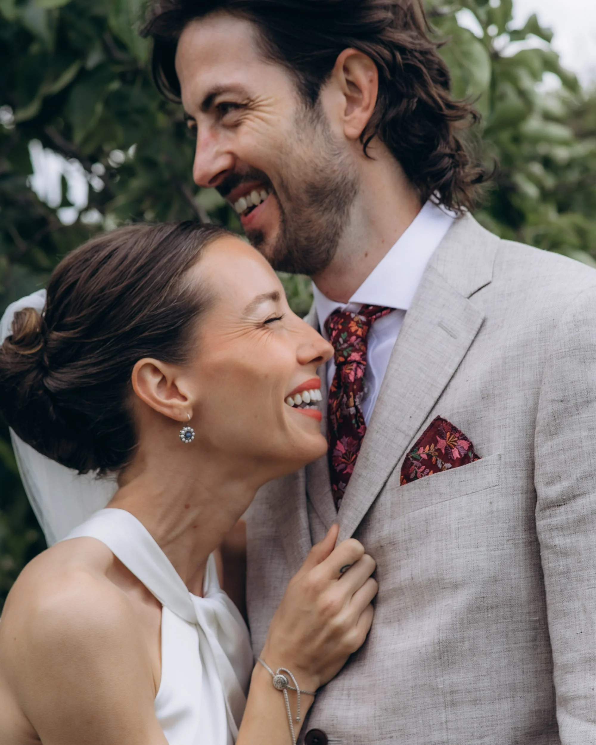  Intimate close-up of the bride and groom laughing together, the bride clutching the groom's lapel, both faces lit with genuine joy, surrounded by soft green foliage 