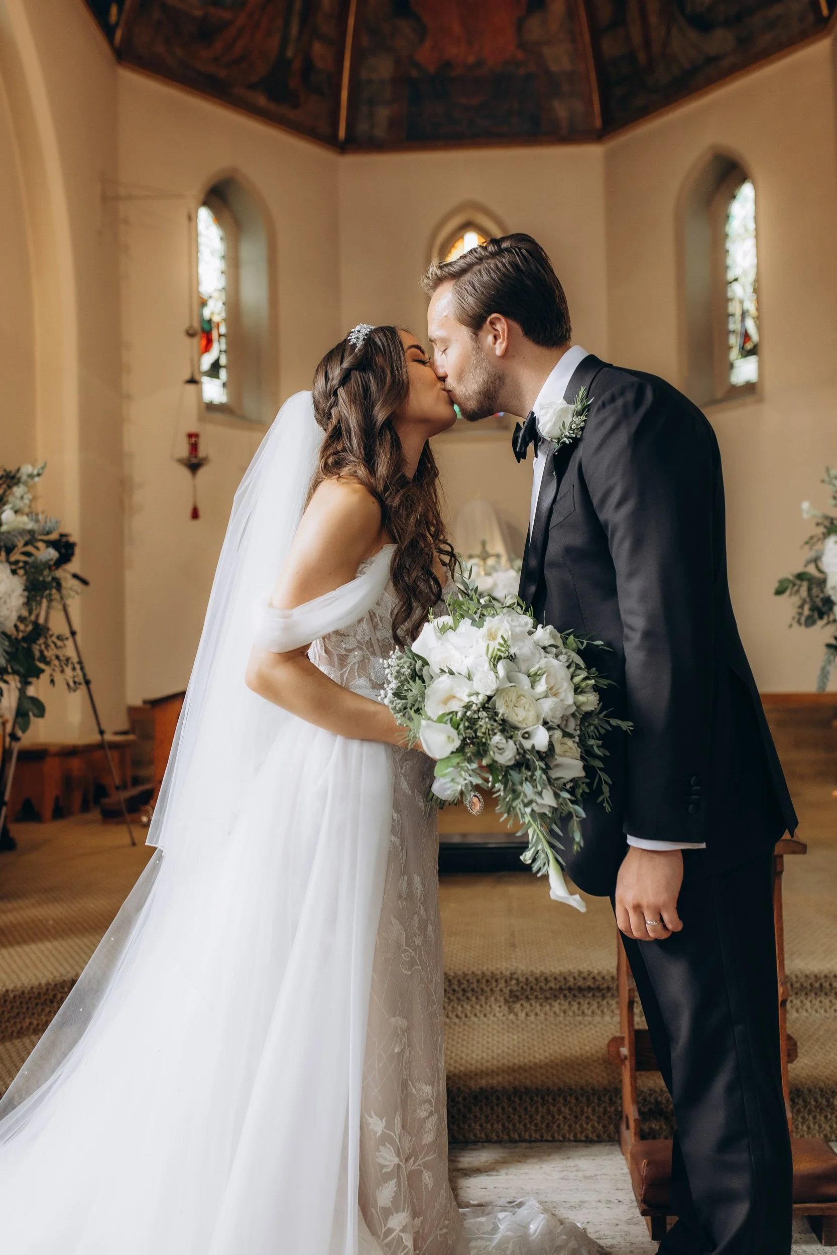 Romantic first kiss of a bride and groom at the altar of a historic London church; timeless wedding photography capturing authentic love by Lusy Klintsova.