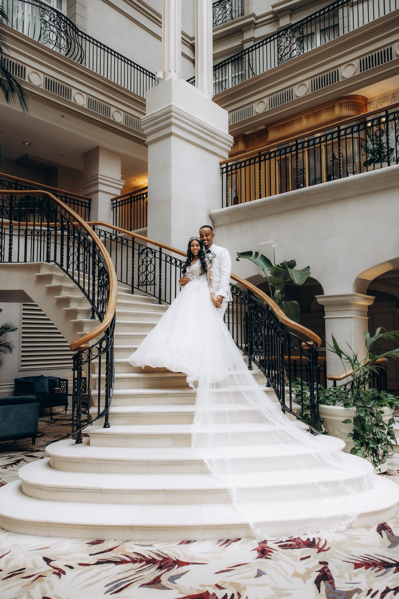 Breathtaking portrait of a smiling bride and groom in an all-white tuxedo posing on the stunning, grand marble staircase of [Specific Luxury London Hotel], showcasing the opulence of the high-ticket venue and expert photography.