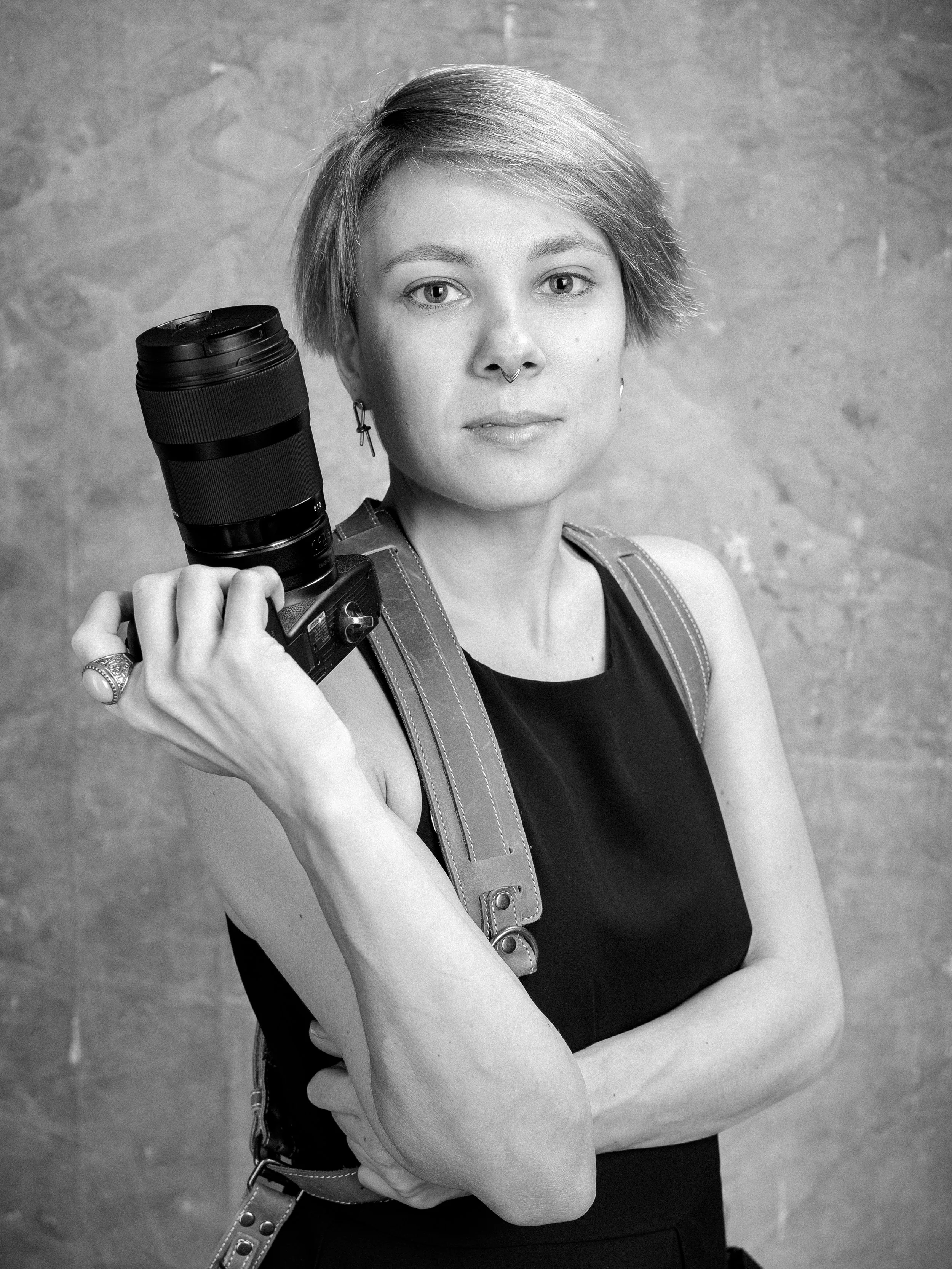 Black and white photo of a woman holding a camera with a telephoto lens resting on her shoulder. She has short hair, a septum piercing, and wears a sleeveless top and earrings. She is standing in front of a plain wall.