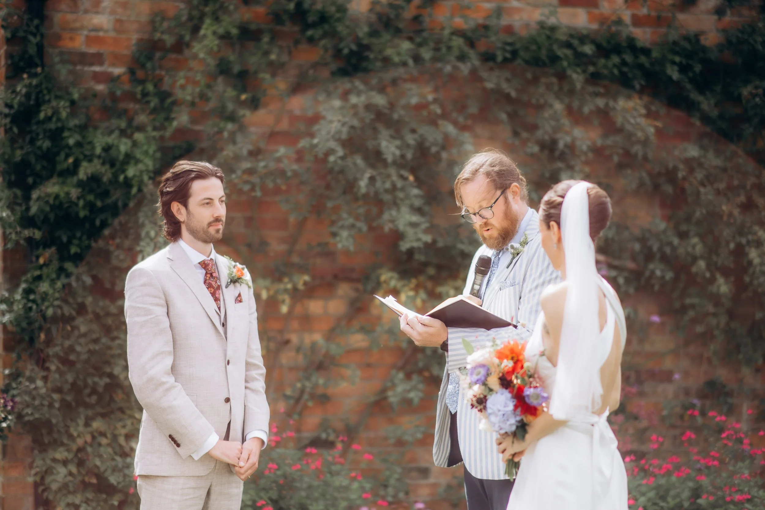 Outdoor ceremony scene against an ivy-covered brick wall, groom in a light grey suit standing composed while the officiant in a striped jacket reads aloud, bride with her back to camera holding a vibrant mixed bouquet
