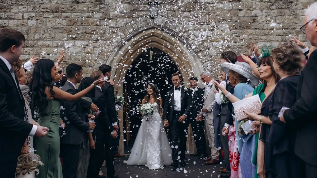 Bride and groom holding hands walking out of a church surrounded by guests throwing confetti.