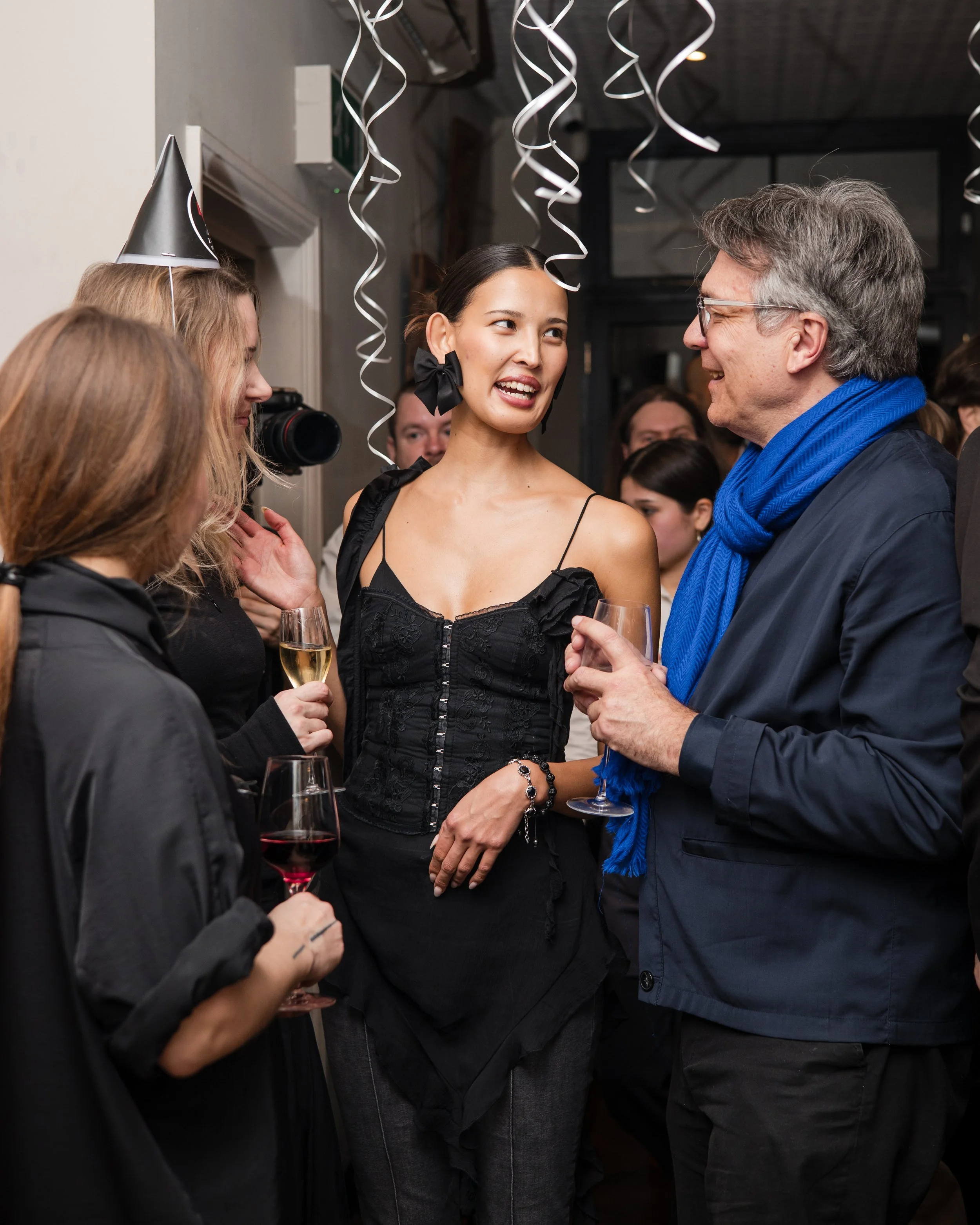 A group of people at a celebration, with two women in the center talking and holding glasses of champagne. One woman has earrings and a black dress, wearing a black bow on her ear. The man on the right is smiling and wearing glasses, a blue scarf, and a dark jacket. Party hats and streamers hang overhead.