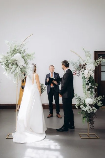 Bride and groom exchanging vows at a floral arch decorated with white and green flowers, officiant facing them in Fulham library