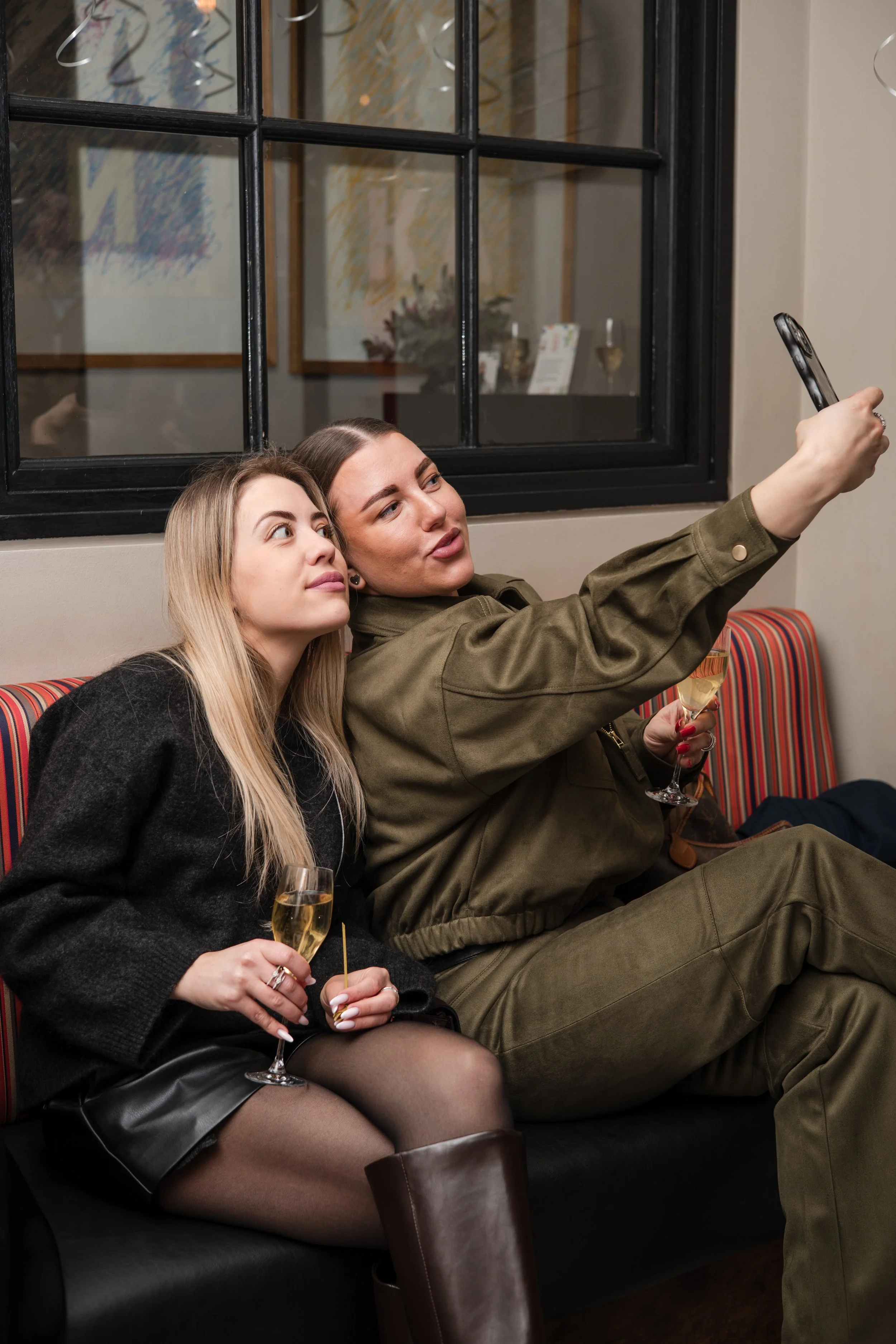 Two women sitting on a black sofa, taking a selfie together. One woman has long blonde hair, wearing a black sweater, and holding a glass of champagne. The other woman has brown hair, dressed in a green outfit, and also holding a glass of champagne. They are inside a room with large window panes and colorful artwork visible in the reflection.