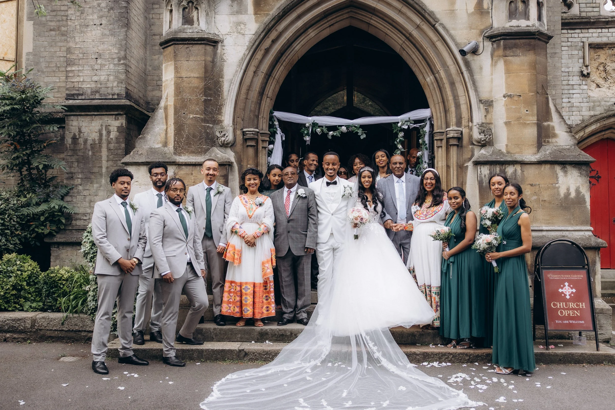 Formal group portrait of the bride, groom, and their large wedding party standing on the steps of a traditional London church venue.