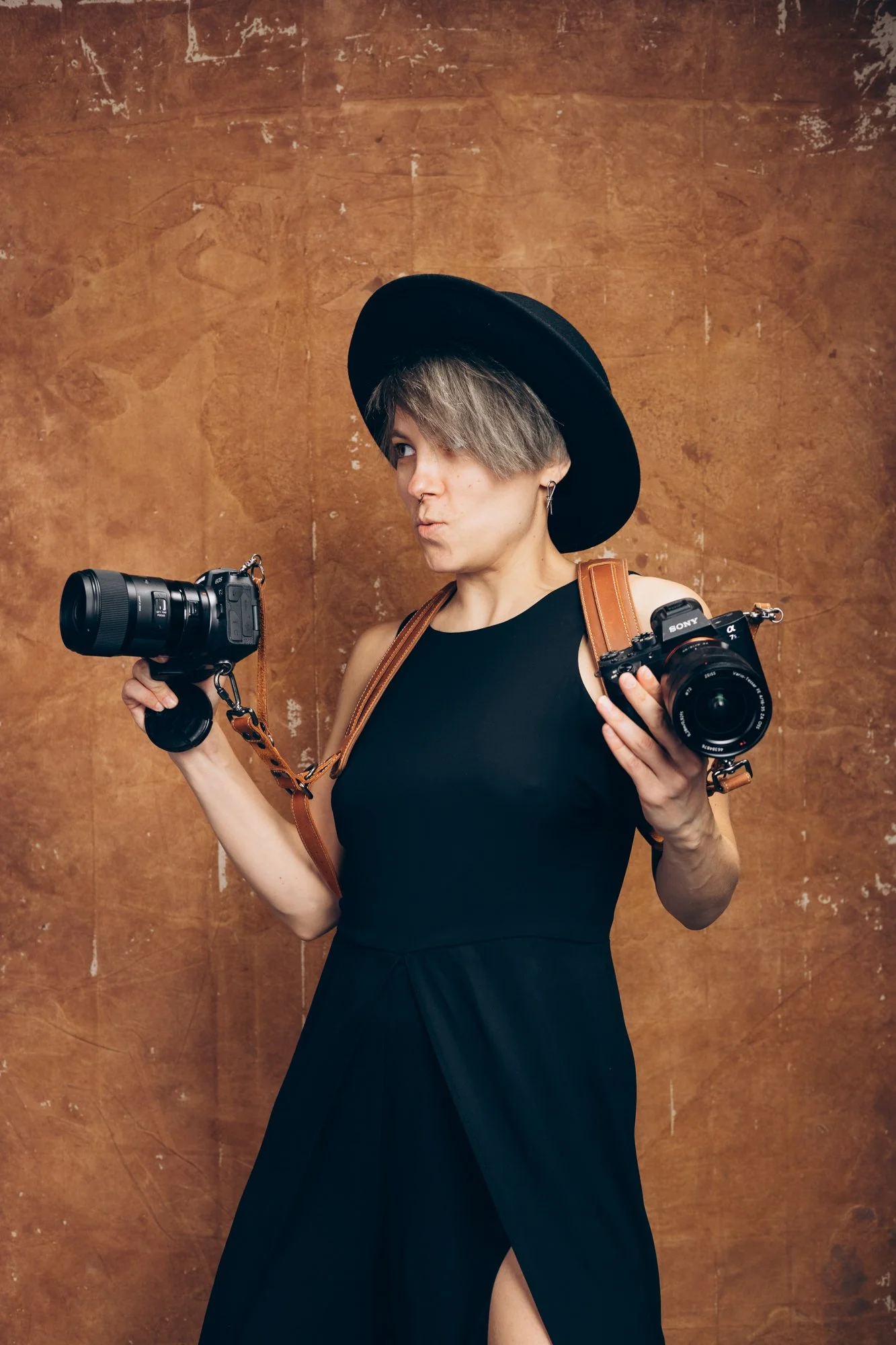 A woman with short gray hair, wearing a black dress with a thigh slit, a wide-brimmed black hat, and earrings, holding a camera in one hand and another camera with a strap over her shoulder, standing against a textured brown background.