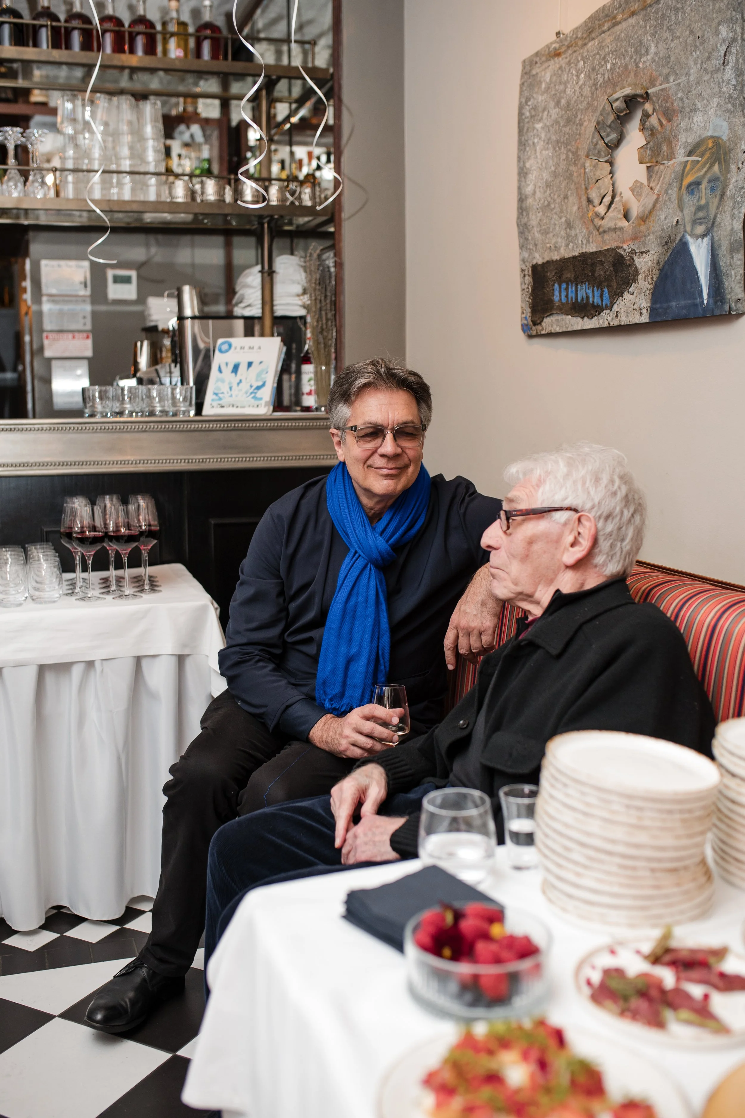 elderly men and a middle-aged man seated around a table at a celebration or gathering. The man is holding a glass of wine, and there are plates of hors d'oeuvres and desserts on the table. The background features a bar with glasses, bottles, and decorations, along with a piece of artwork on the wall.