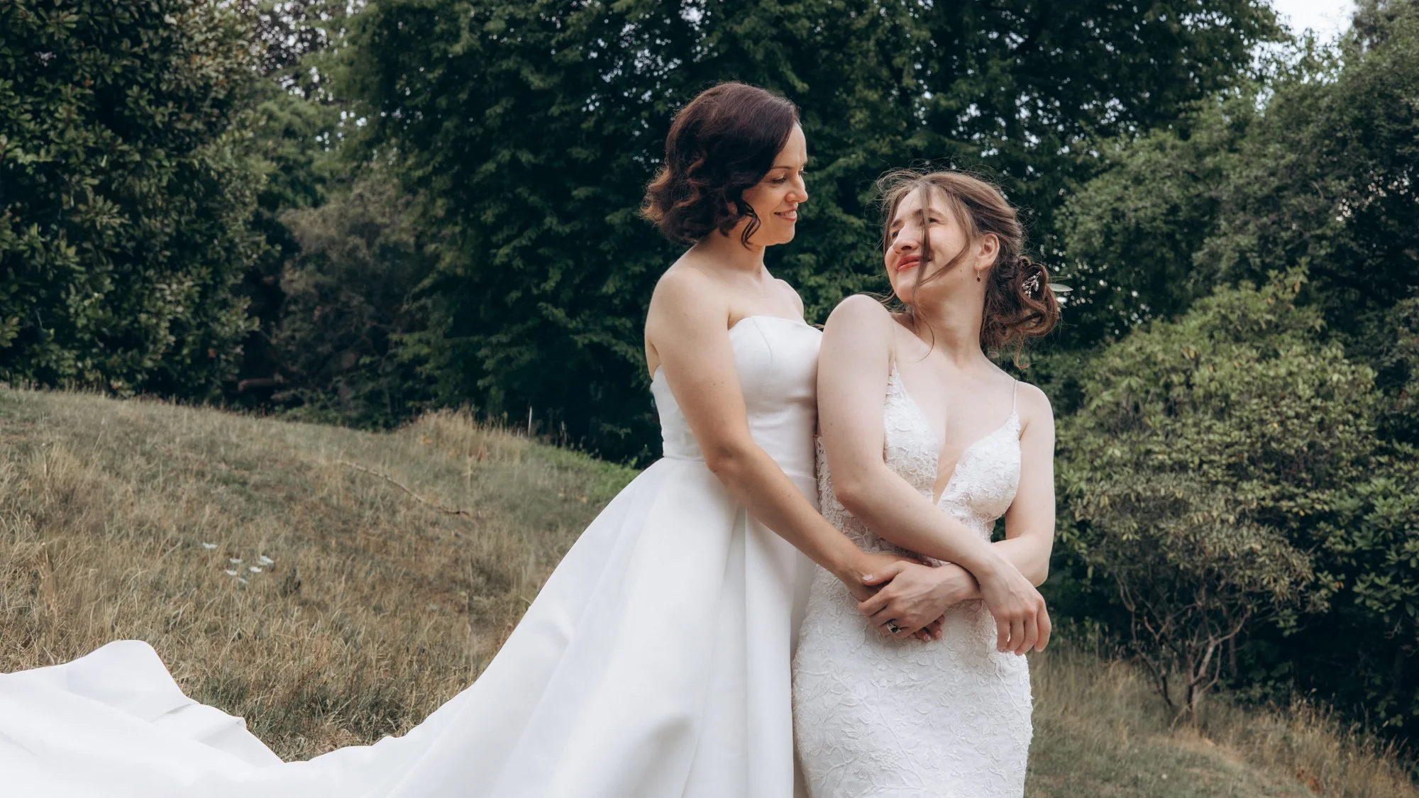 Two women in wedding dresses stand outdoors in a grassy area with trees in the background, embracing and smiling.