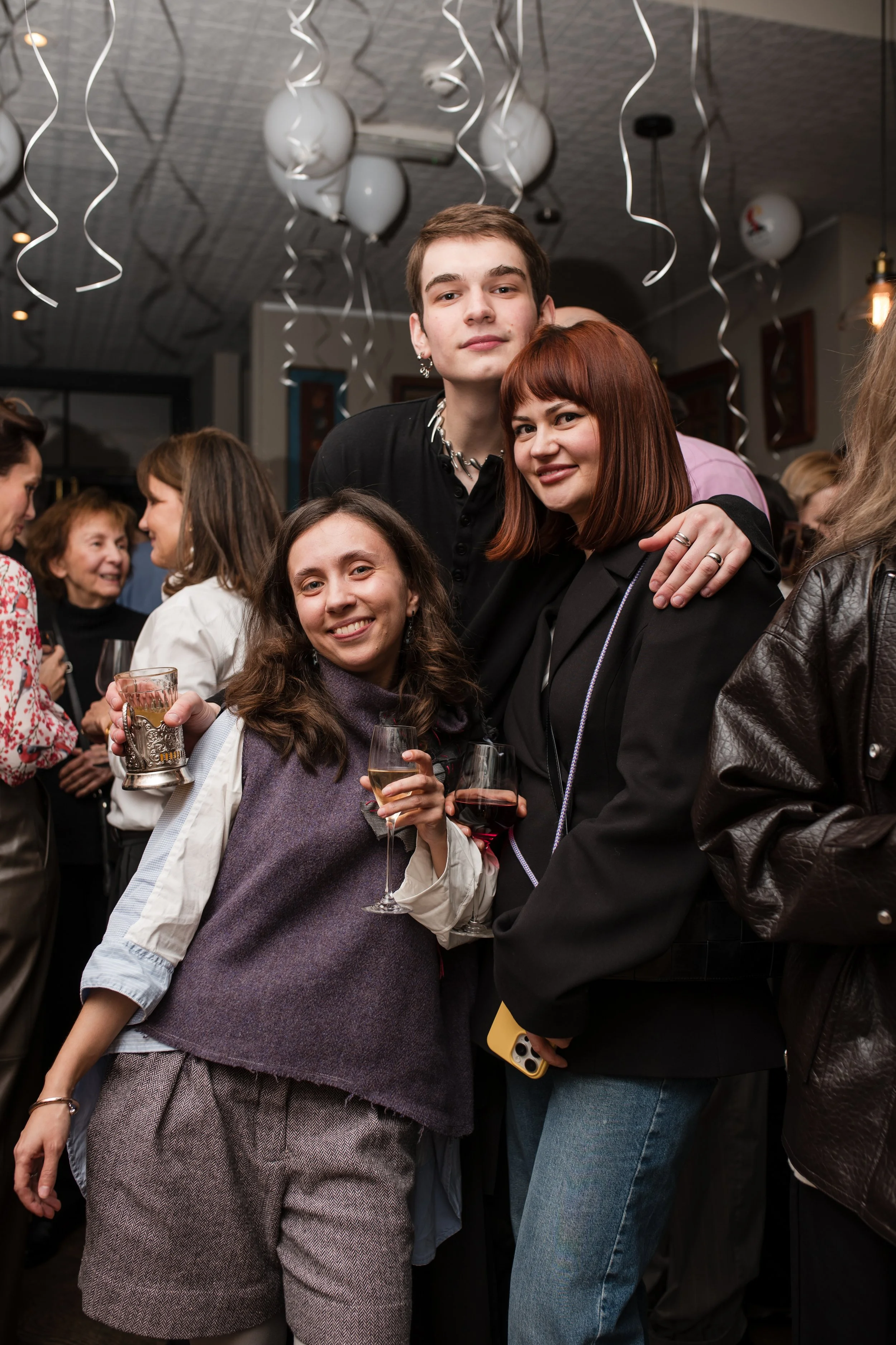 Group of young people at a celebration, holding drinks and smiling, with white balloons and hanging streamers in the background.