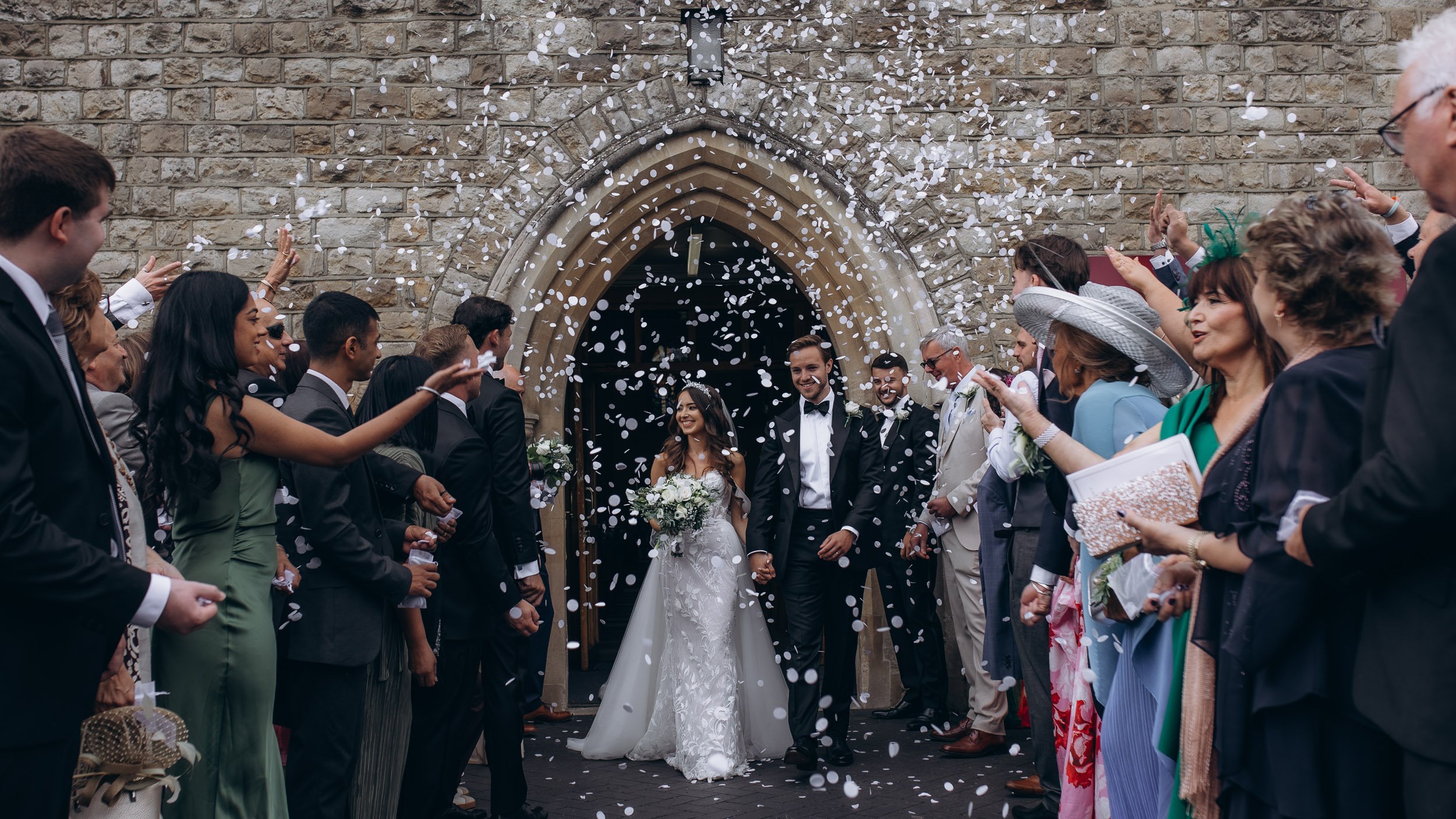 A bride and groom walking out of a church while guests throw confetti. The church has a stone facade and an arched doorway. The bride is holding a bouquet and wearing a white wedding dress, and the groom is in a dark suit with a bow tie. Guests are dressed in formal attire, some wearing hats, and are celebrating as they watch the couple.