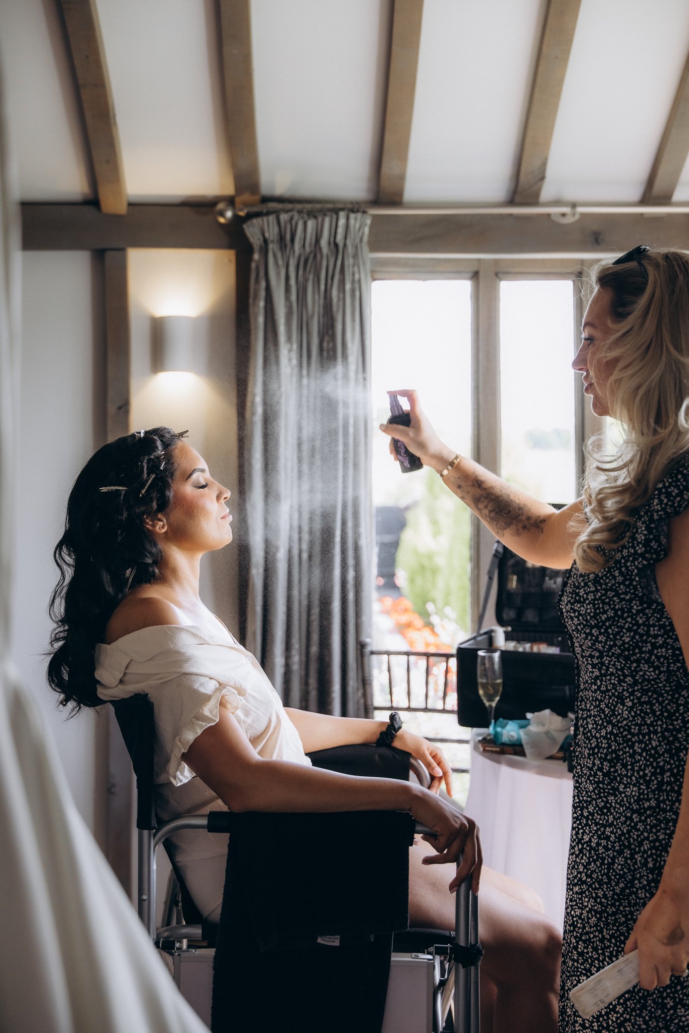 Hairdresser spraying a bride's curled hair while a guest photographs on her phone