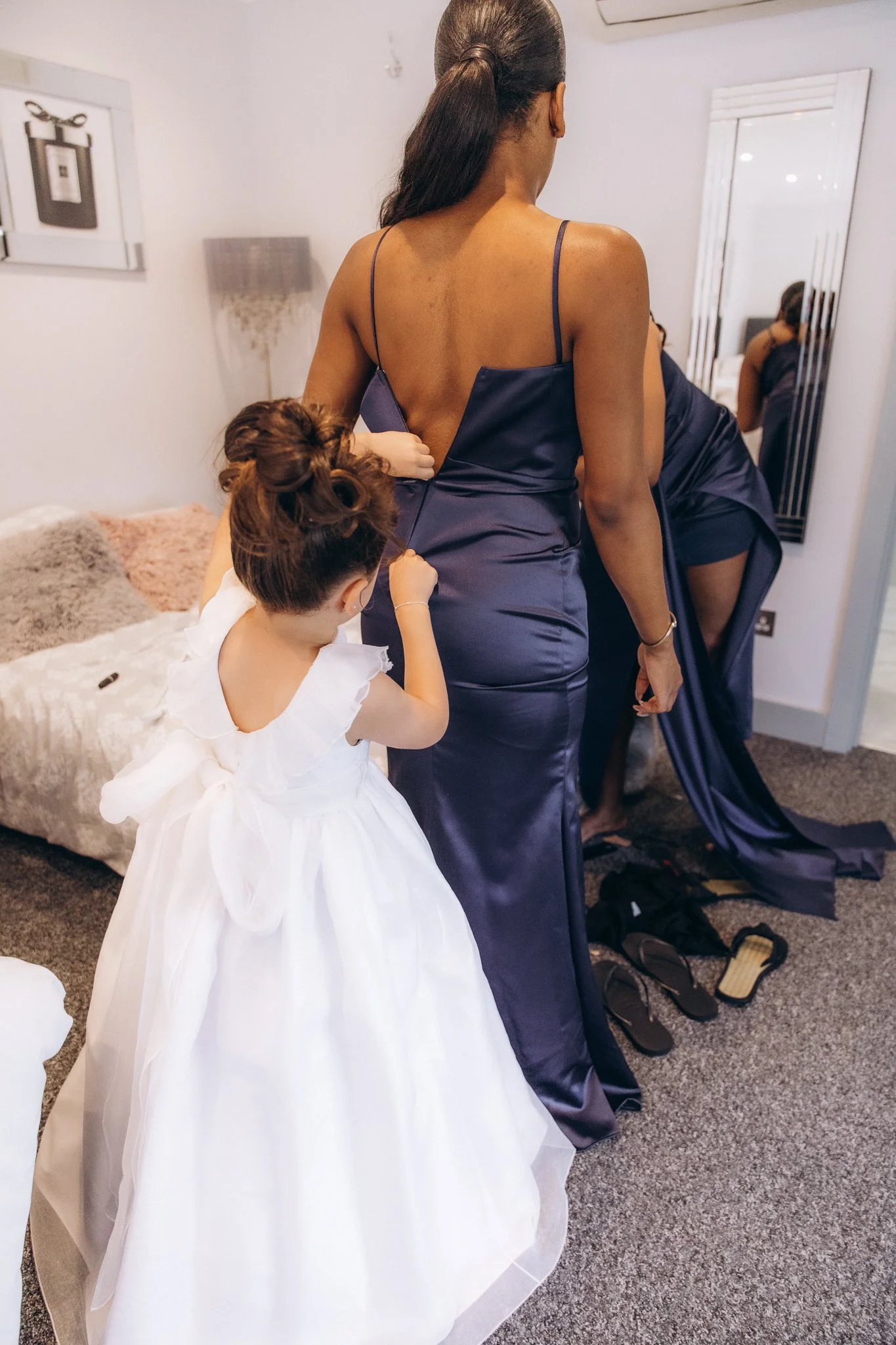 Flower girl having her white dress fitted by a bridesmaid in a navy gown